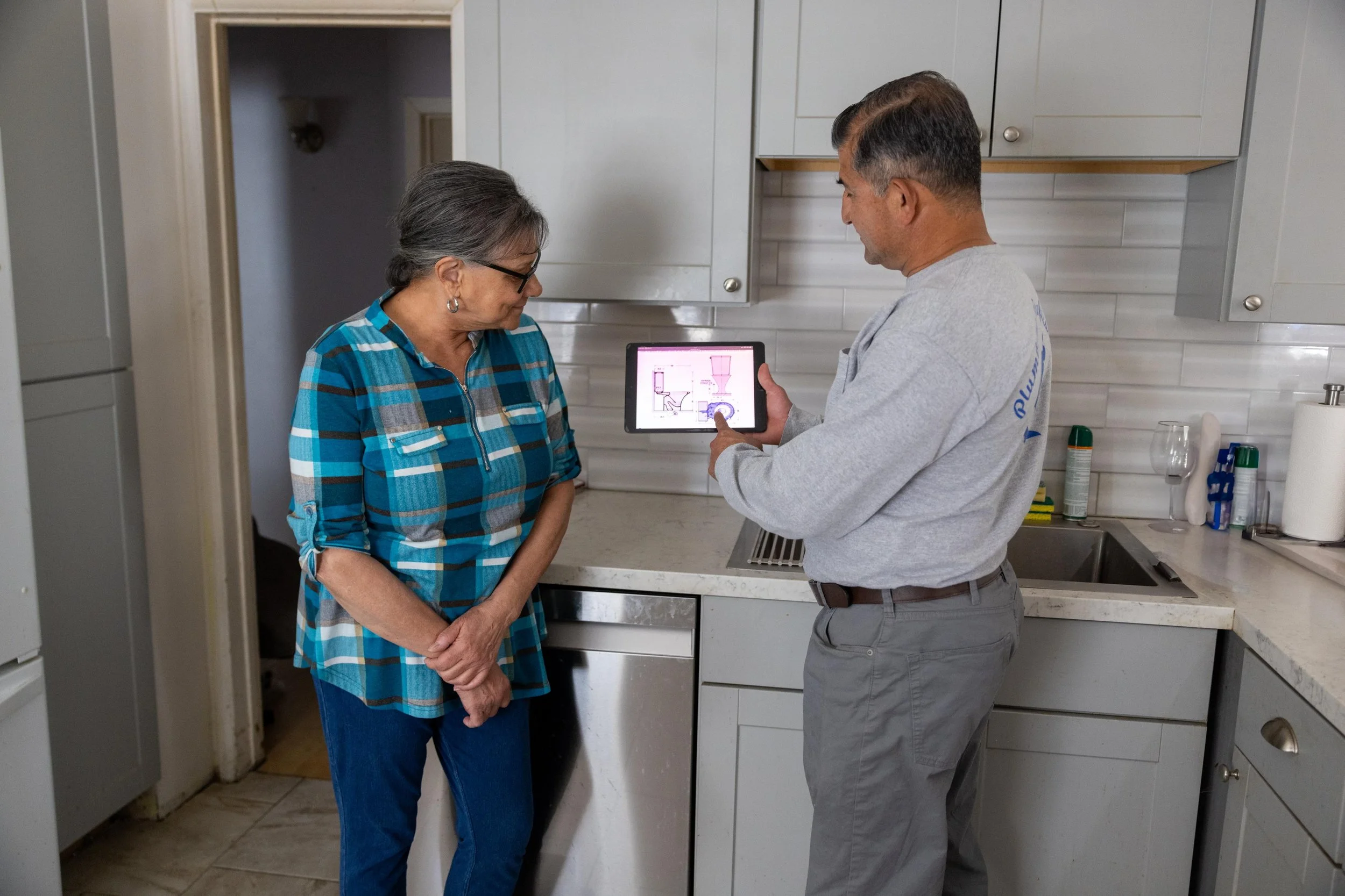 A man and woman in a kitchen looking at a tablet displaying a diagram of a toilet. The woman is wearing glasses and a blue checkered shirt, while the man is wearing a gray shirt and gray pants.