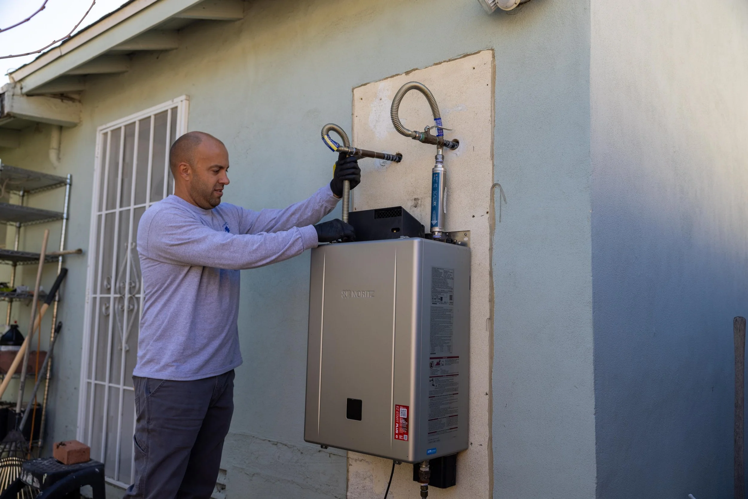 A man installing or maintaining outdoor water or gas lines on a house wall with two flexible hoses connected to pipes above a tankless water heater