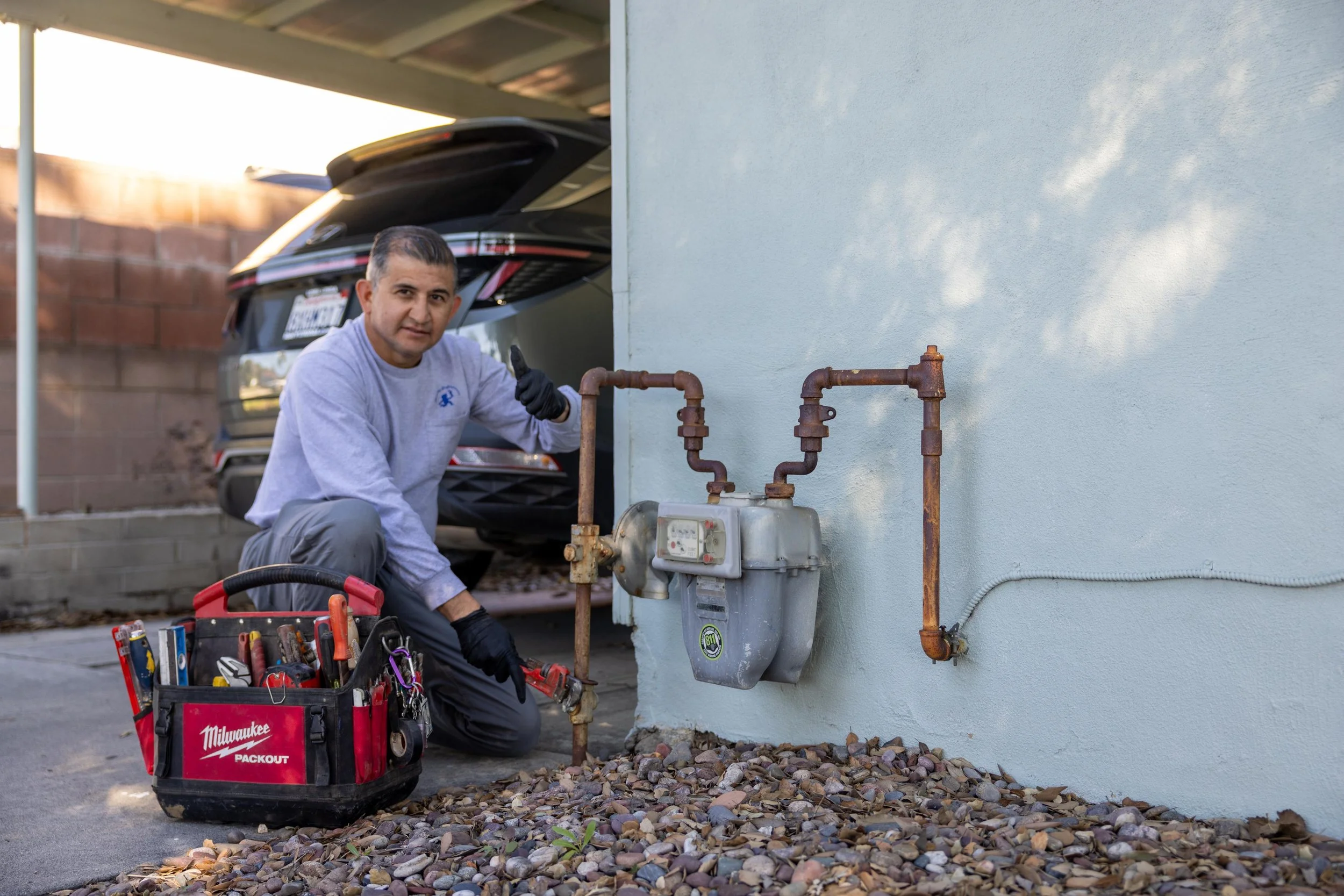A man working on plumbing outside a house, kneeling next to a utility box with tools in a red Milwaukee toolbox, with a black SUV parked under a carport behind him.