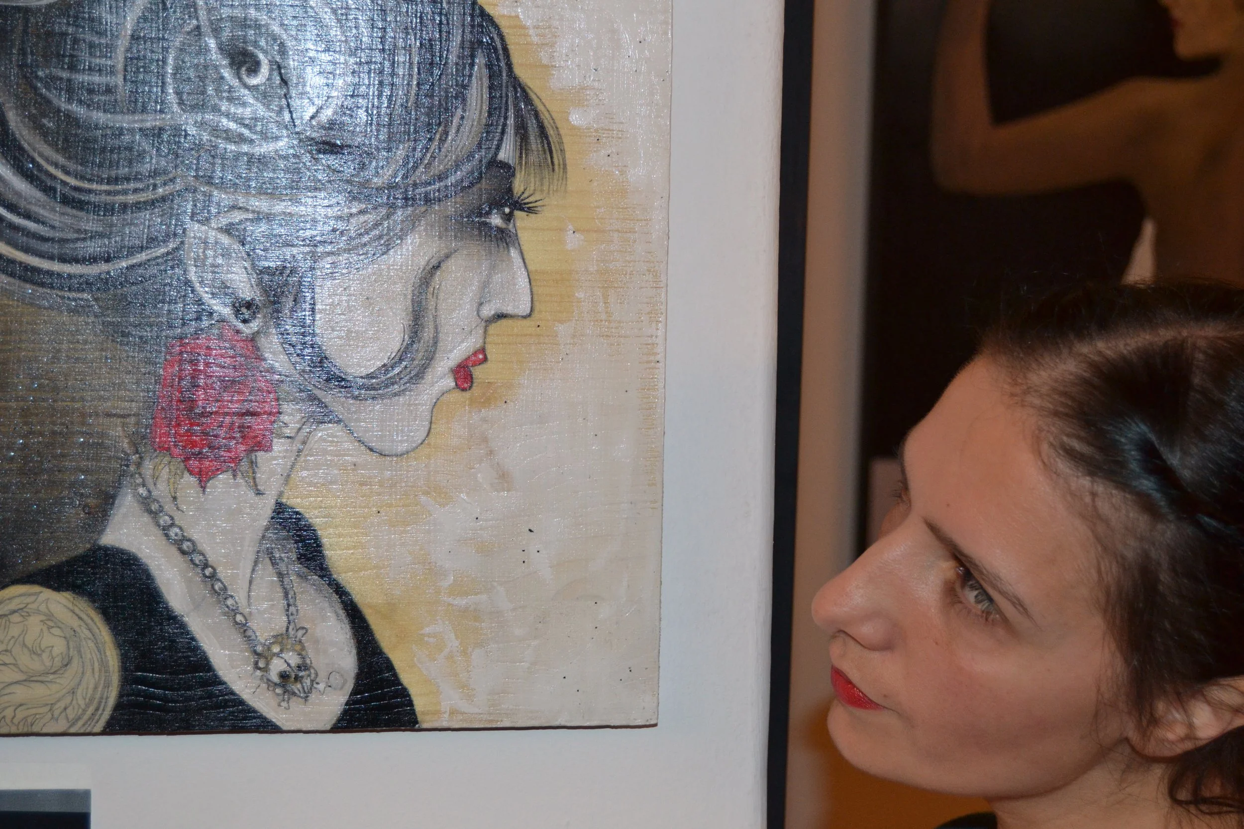 Una giovane donna osserva un dipinto sulla parete, raffigurante un ritratto stilizzato di una donna con capelli grigi, rossetto rosso e collana, con un orecchino rosso.