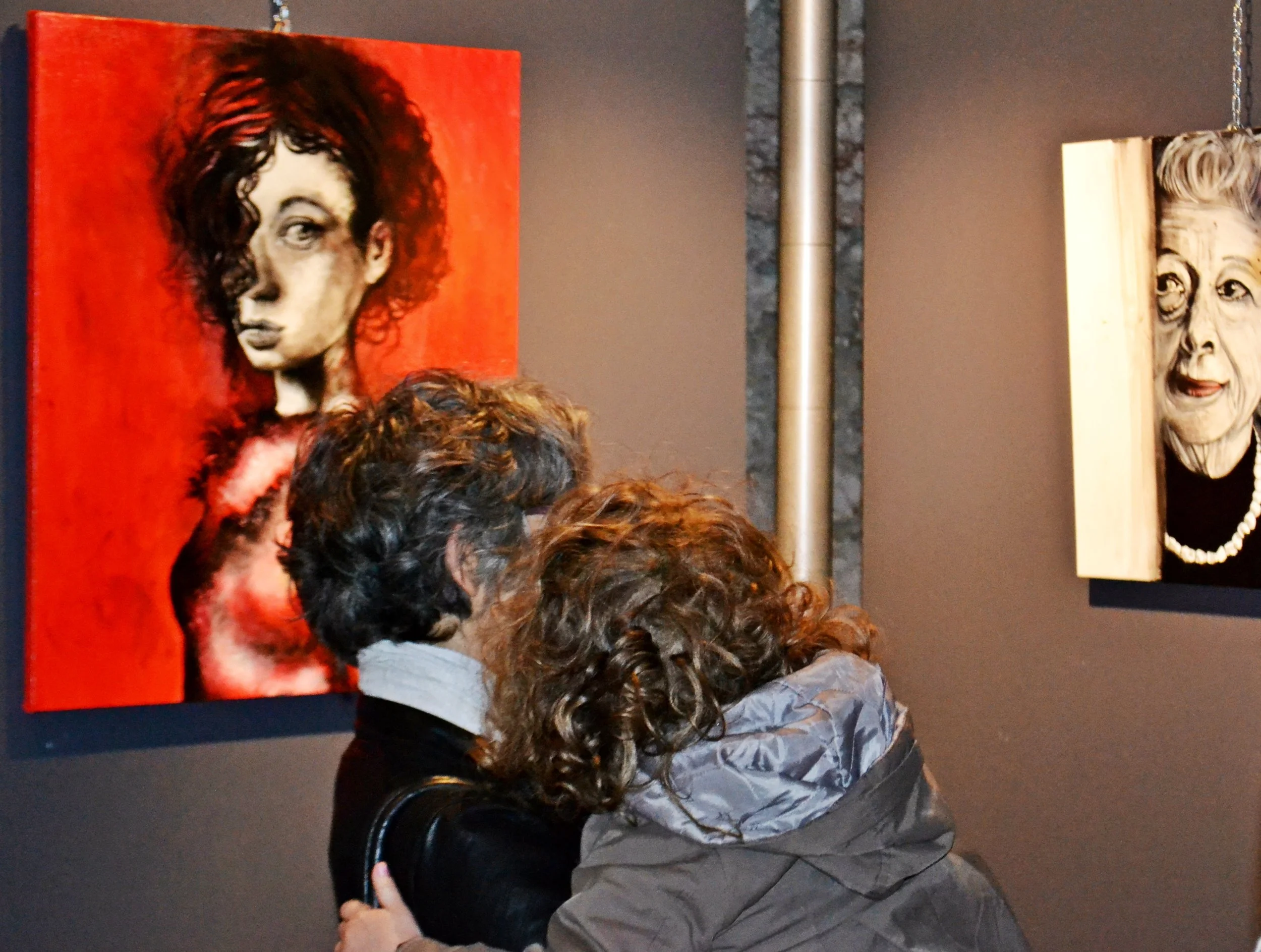 Due persone che osservano quadri in una mostra d'arte. Uno dei quadri raffigura un volto femminile con abbigliamento di colore nero e una collana di perle, l'altro è un ritratto di una donna anziana con capelli gray e occhi espressivi.
