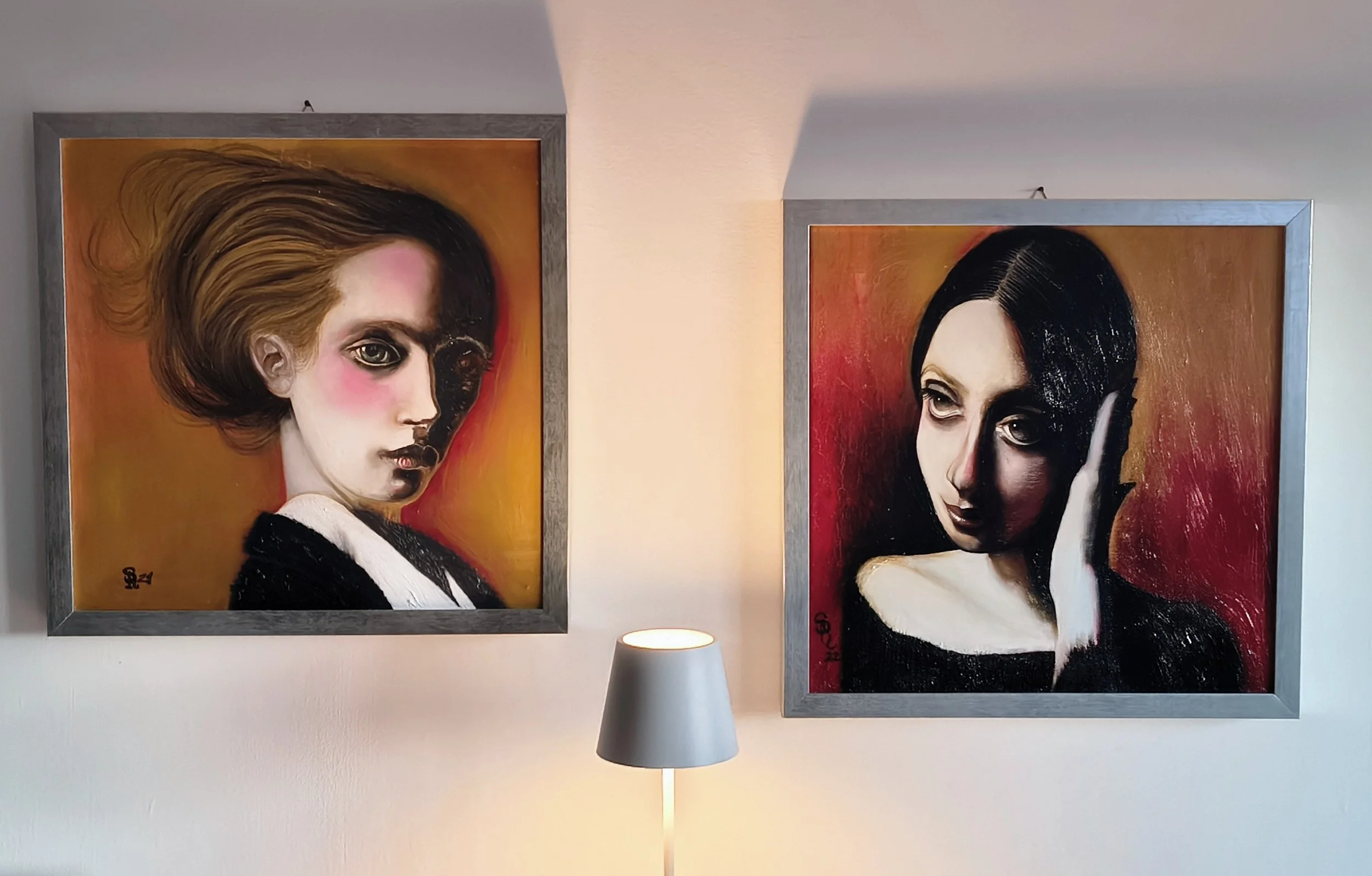 Due dipinti di ritratti femminili moderni con sfondi arancioni e rossi, incorniciati in cornici di legno, appesi a una parete bianca. Un lampada da tavolo con paralume bianco si trova sotto i quadri. Esempio di scelta d'arredo di qualità