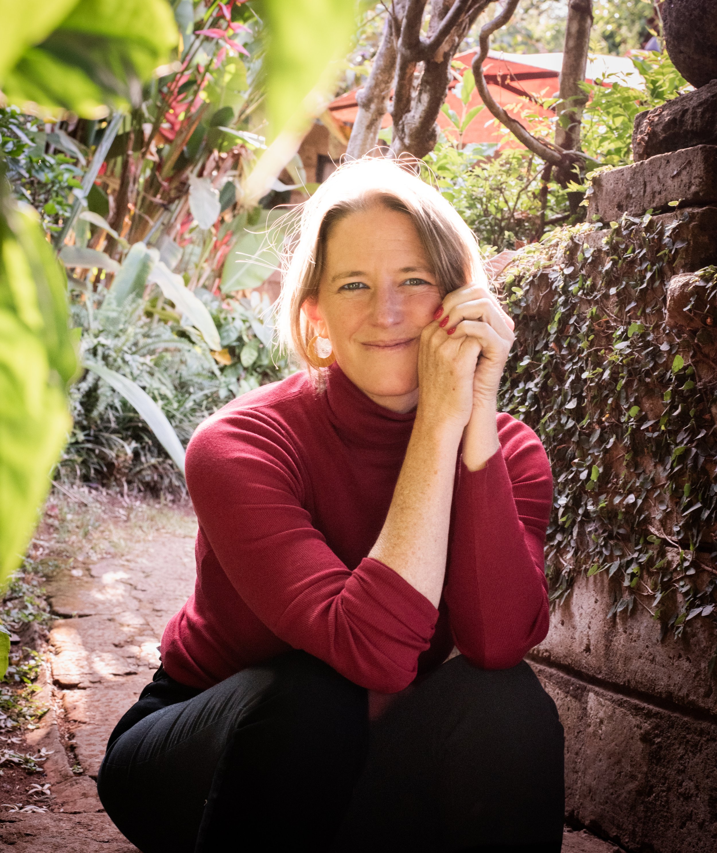 A woman with short hair wearing a red turtleneck sweater, sitting outdoors among greenery, smiling with her hands gently pressed to her cheek.