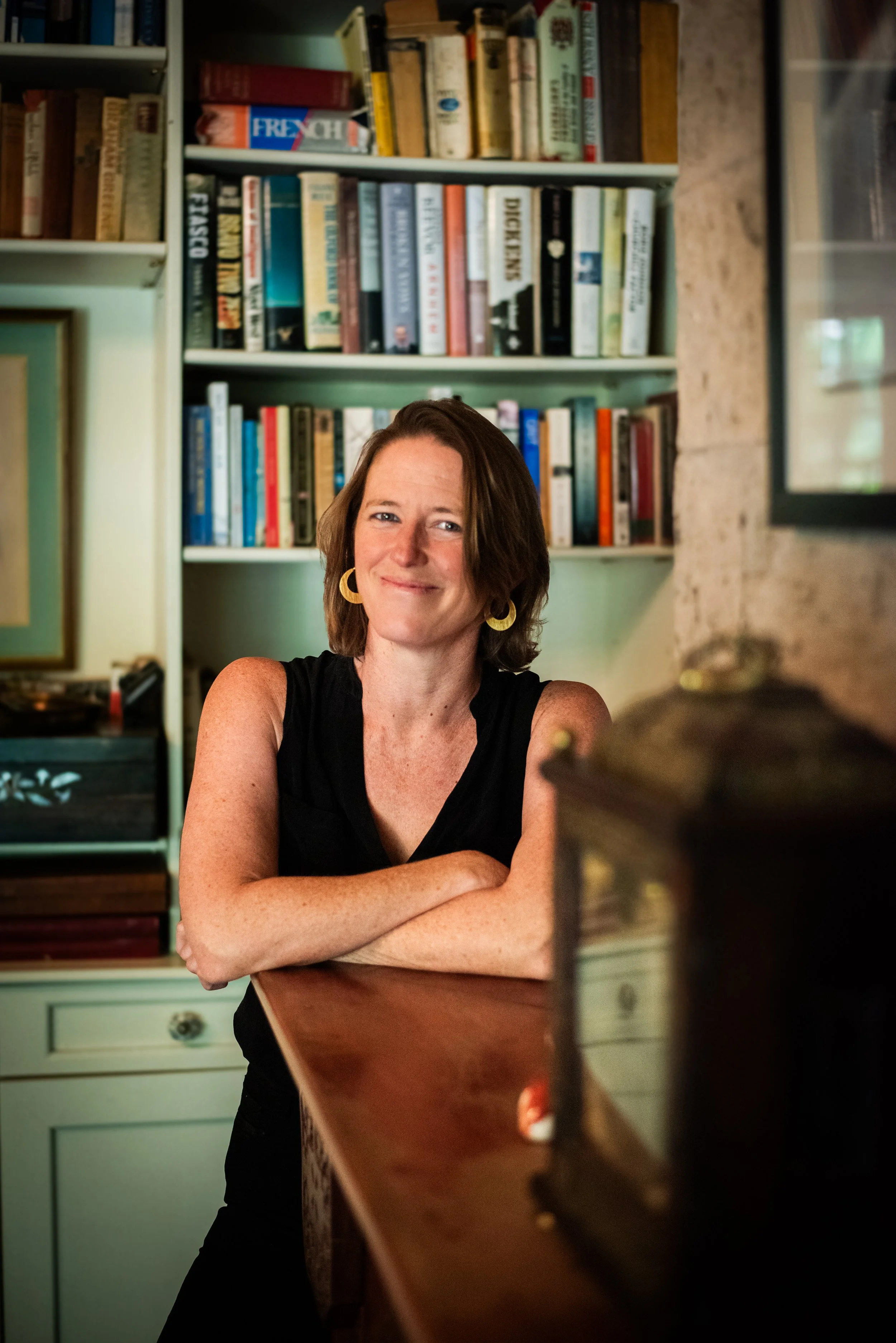A woman with short brown hair and yellow earrings sitting at a wooden table in a room with a bookshelf filled with books in the background.