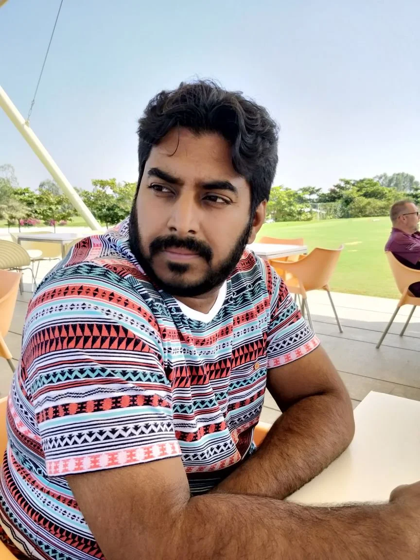 A young man with dark hair and a beard sitting outdoors at a table, wearing a colorful patterned shirt, with a park-like background and a man sitting in the background.