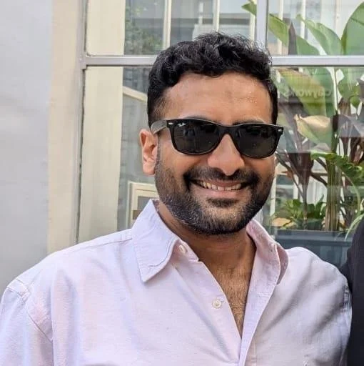 A man with dark curly hair, a beard, and sunglasses smiling, wearing a light pink button-up shirt. He is outdoors with a plant and window in the background.