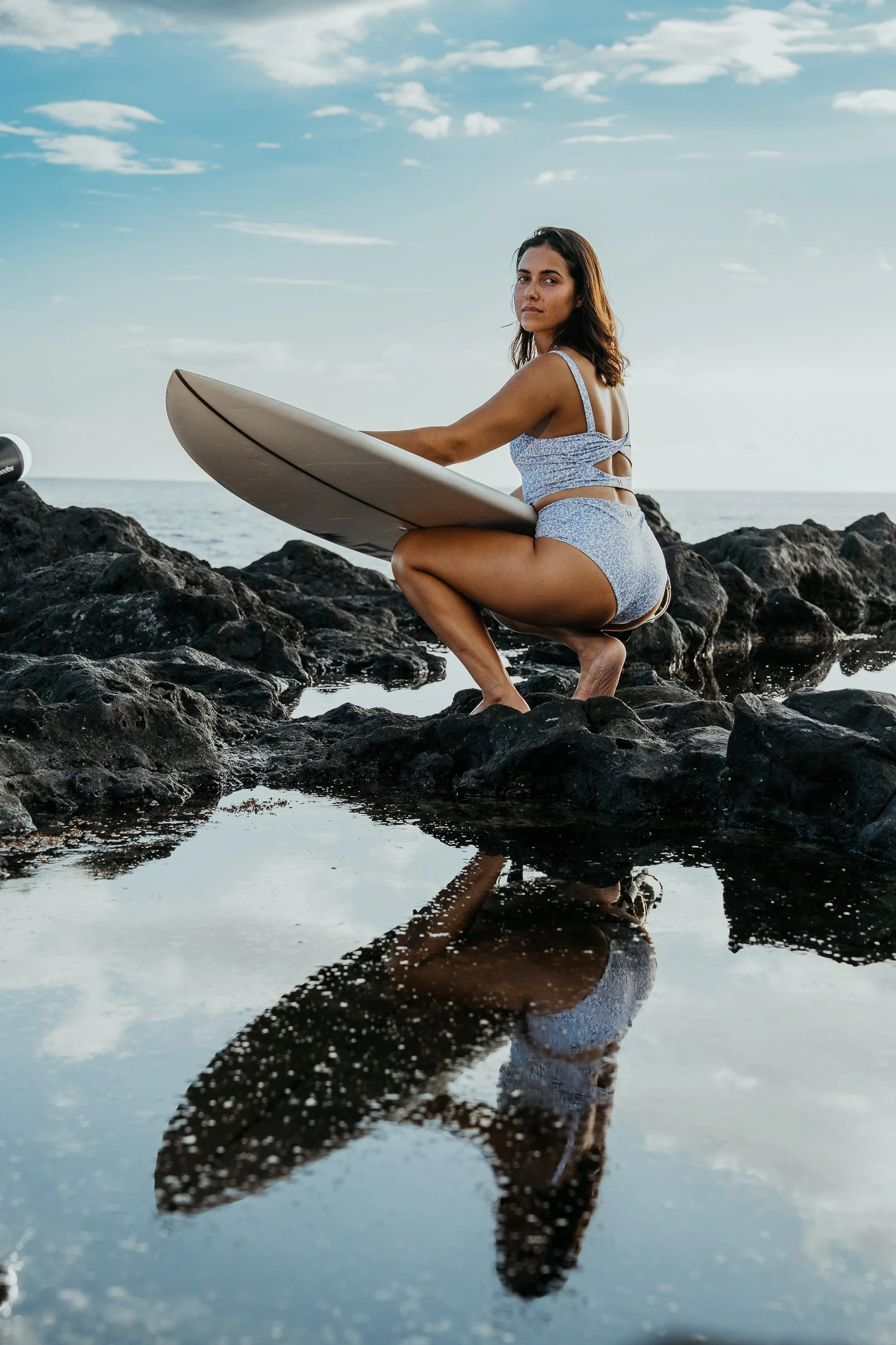 Femme brune en maillot de bain à motifs bleus, tenant une planche de surf, assise sur des rochers au bord de la mer sous un ciel partiellement nuageux.
