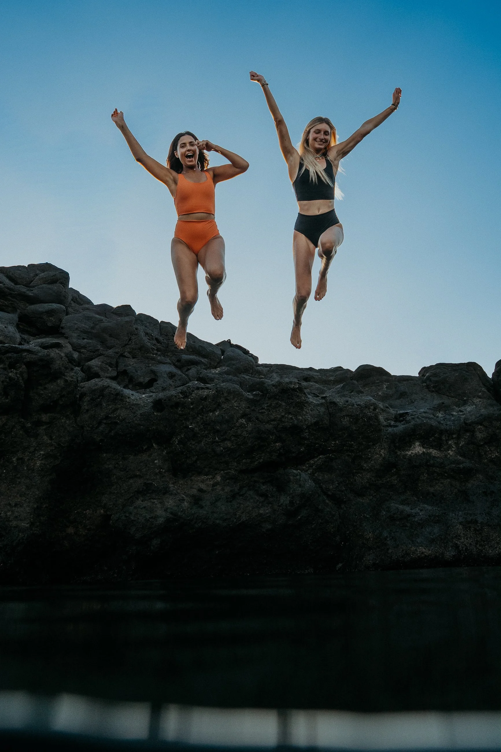 Deux femmes sautent joyeusement sur des rochers au coucher du soleil, avec un ciel clair en arrière-plan.