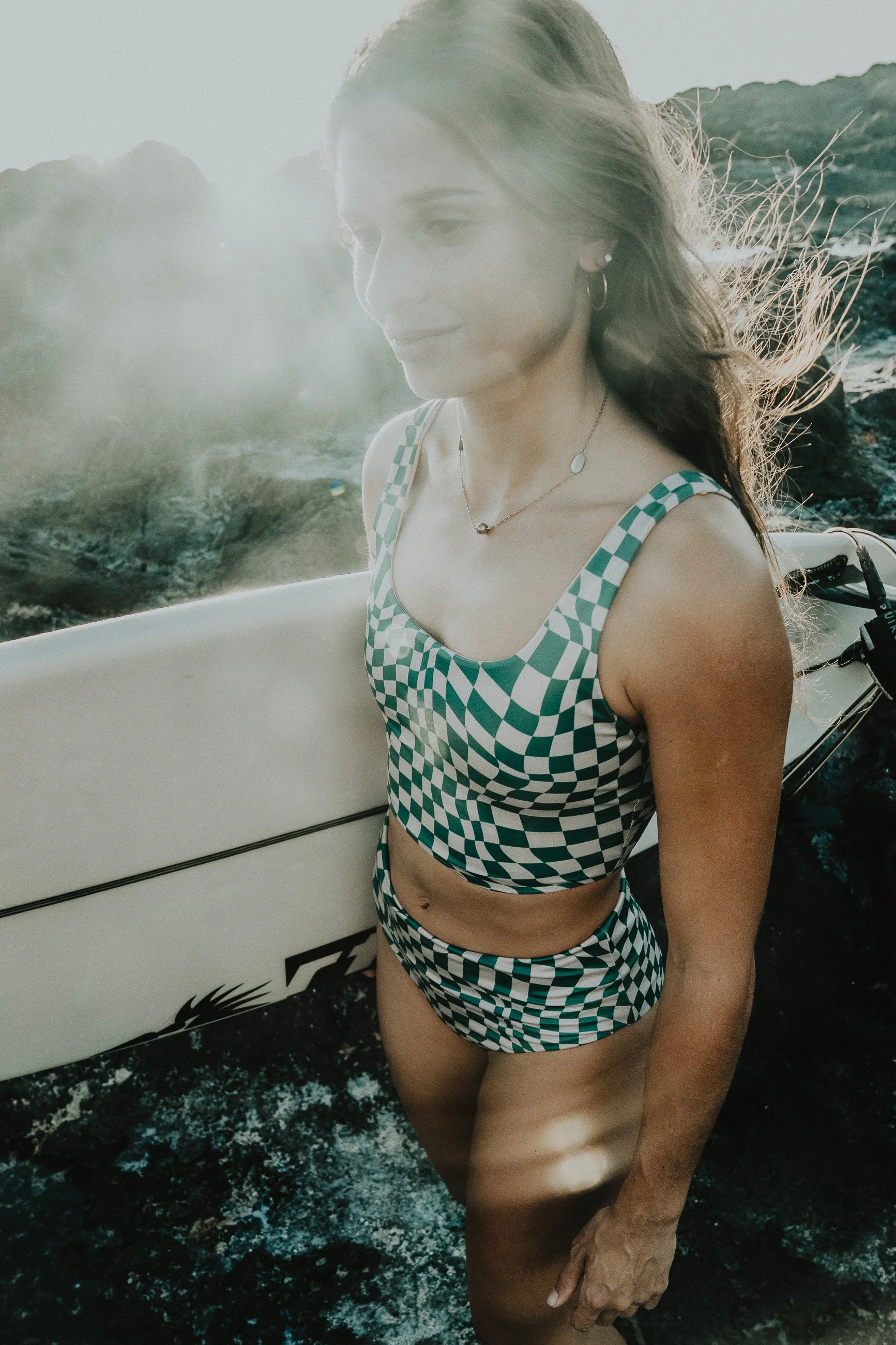 Une femme portant un maillot de bain à carreaux vert et blanc, se tenant devant une planche de surf, avec une mer en arrière-plan et le soleil brillant dans le ciel.