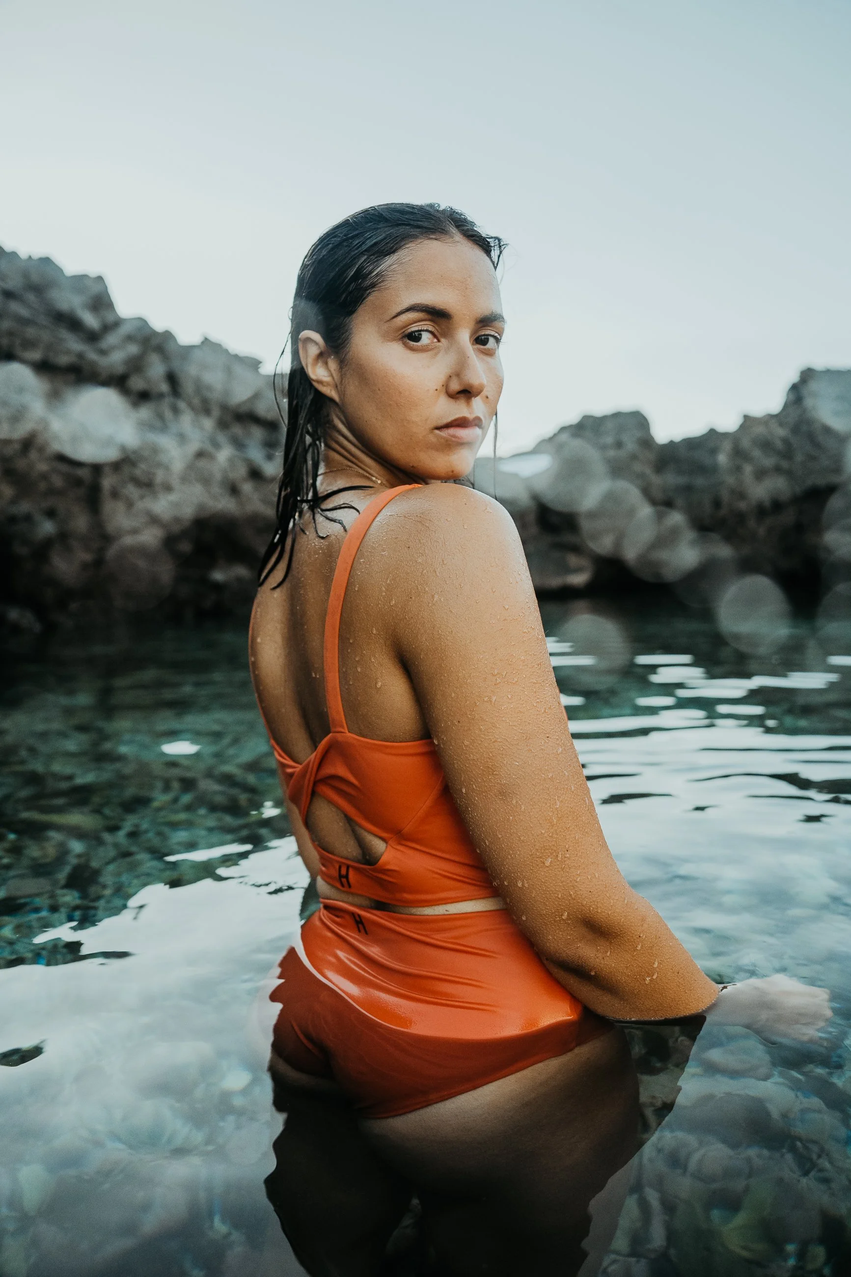 Femme en maillot orange dans l'eau près de rochers, regardant par-dessus l'épaule, avec des rochers en arrière-plan et un ciel clair.