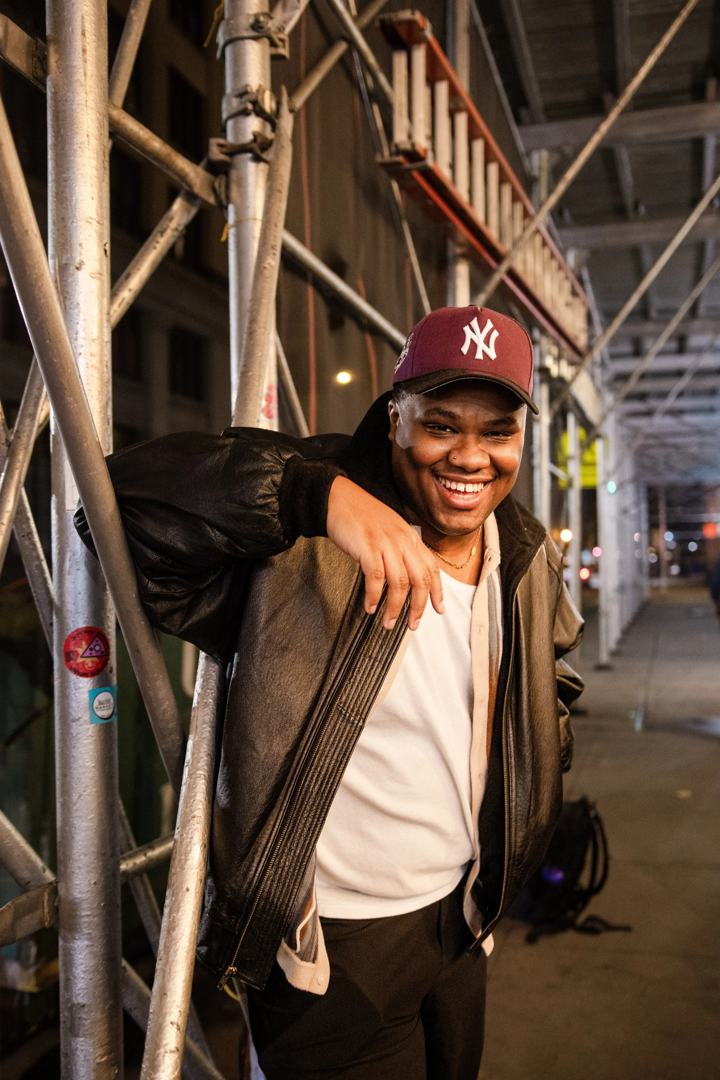 A young man smiling and posing against a metal construction scaffold on a city street at night, wearing a maroon New York Yankees cap and a brown leather jacket.