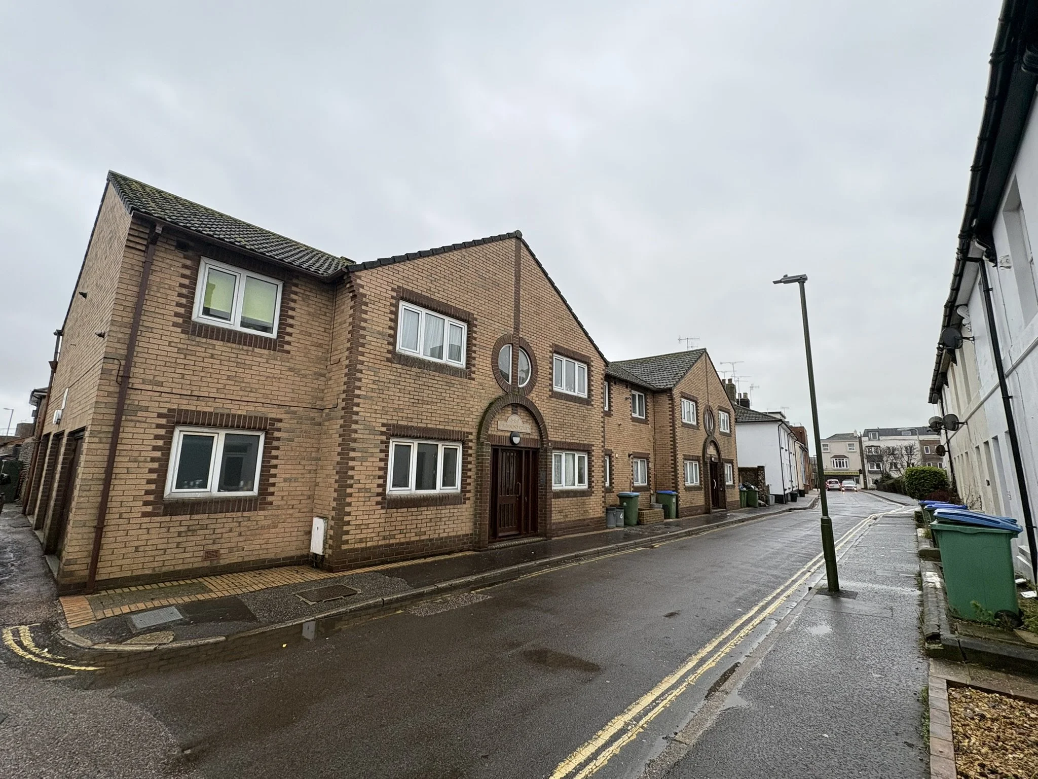 A row of multi-story brick residential buildings on a wet, overcast day, with trash bins on the sidewalk and a street lamp nearby.