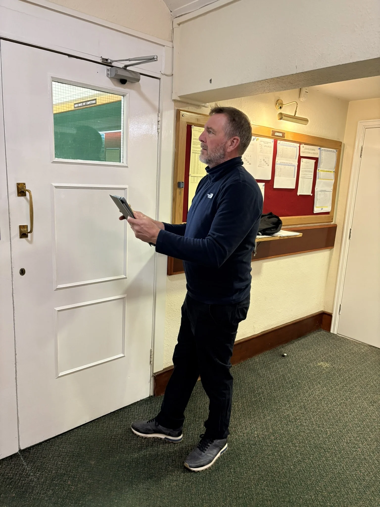 A man standing indoors near a door, holding a tablet, assessing a fire door.