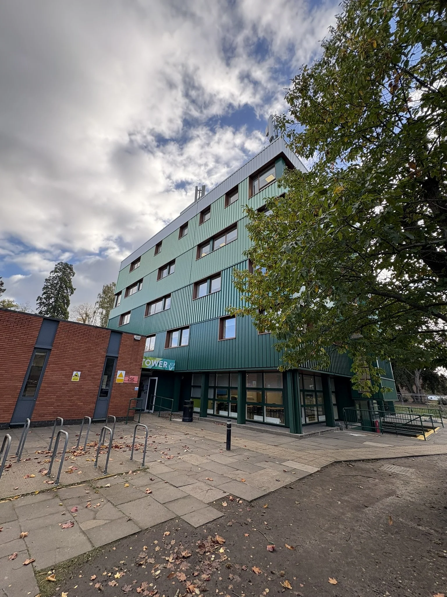 A modern multi-story building with green exterior panels and several windows, surrounded by an outdoor area with paved walkways, bike racks, and a tree with green leaves. The sky above is partly cloudy.