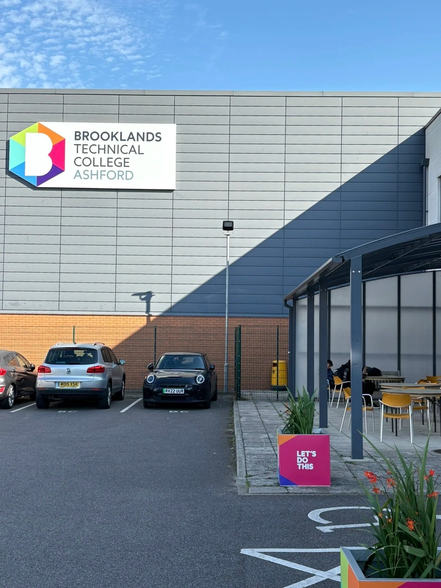Parking lot outside Brooklands Technical College Ashford with a large rectangular sign on the building, several parked cars, and a small seating area with a sign that reads "Let's Do This."