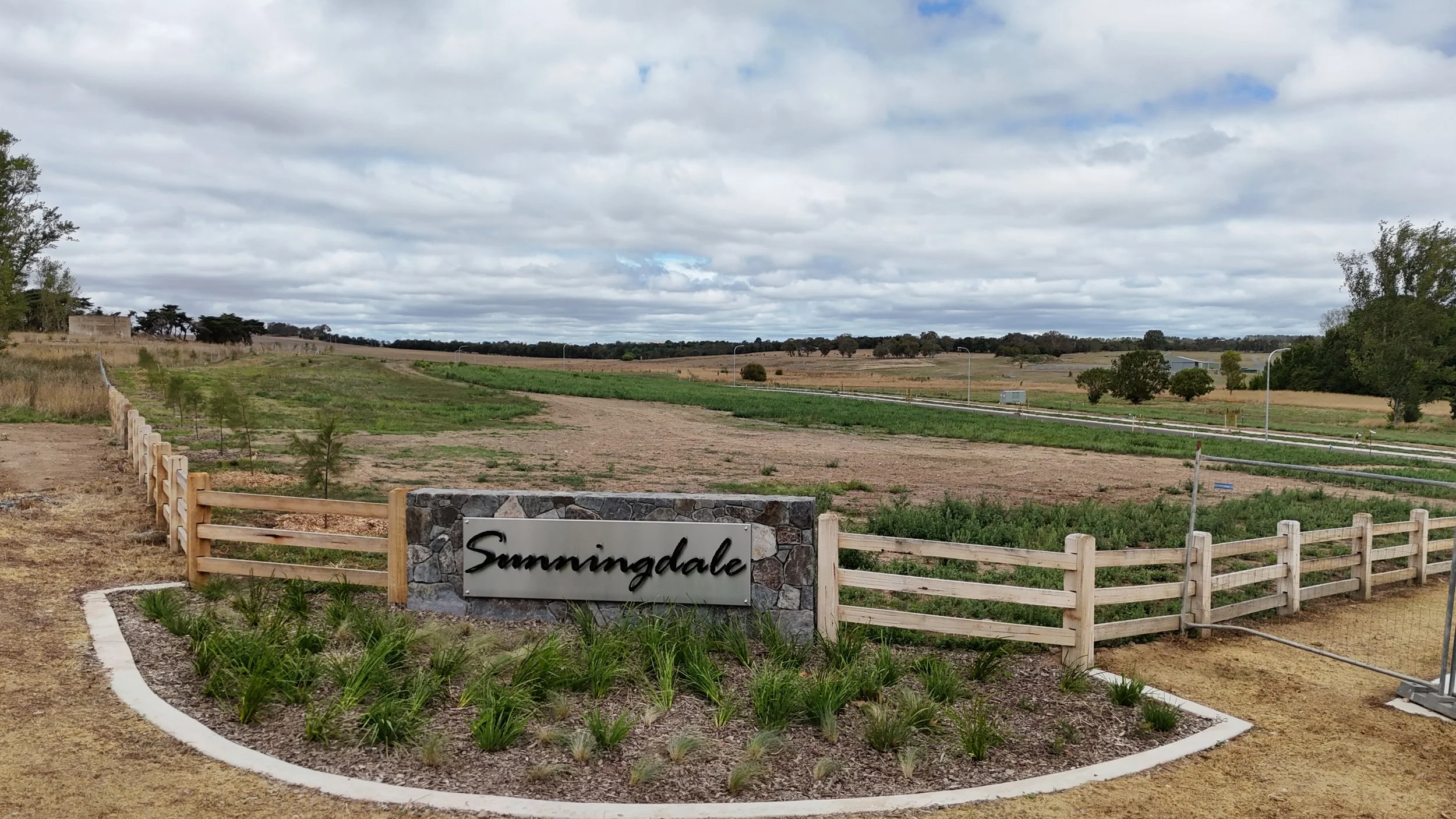 A sign with the word "Summingdale" in front of a landscaped area with plants and a wooden fence, overlooking expansive fields, a road, and a cloudy sky.