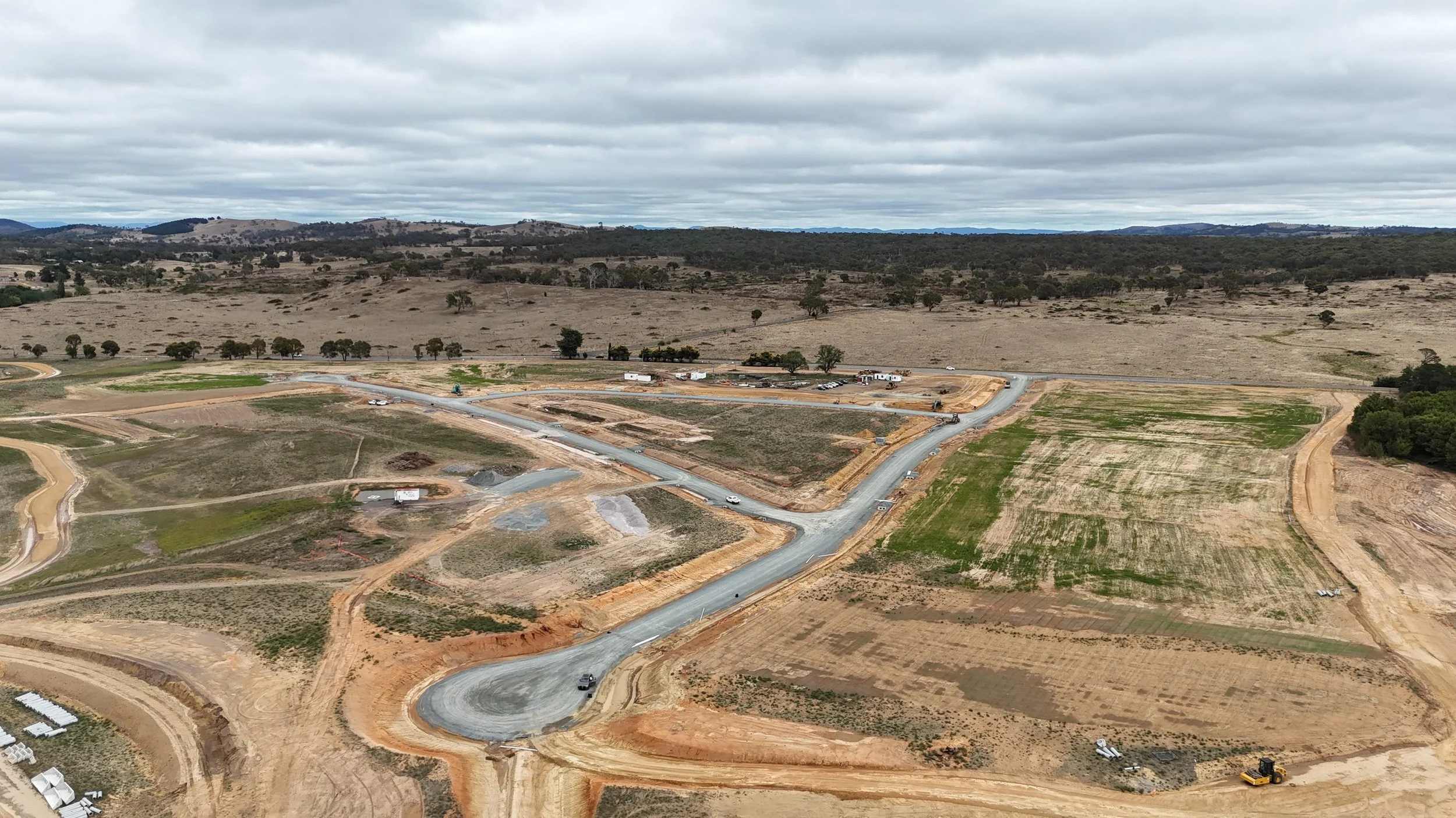 Aerial view of a construction site with roads being built, patches of green grass, and surrounding dry land under a cloudy sky.