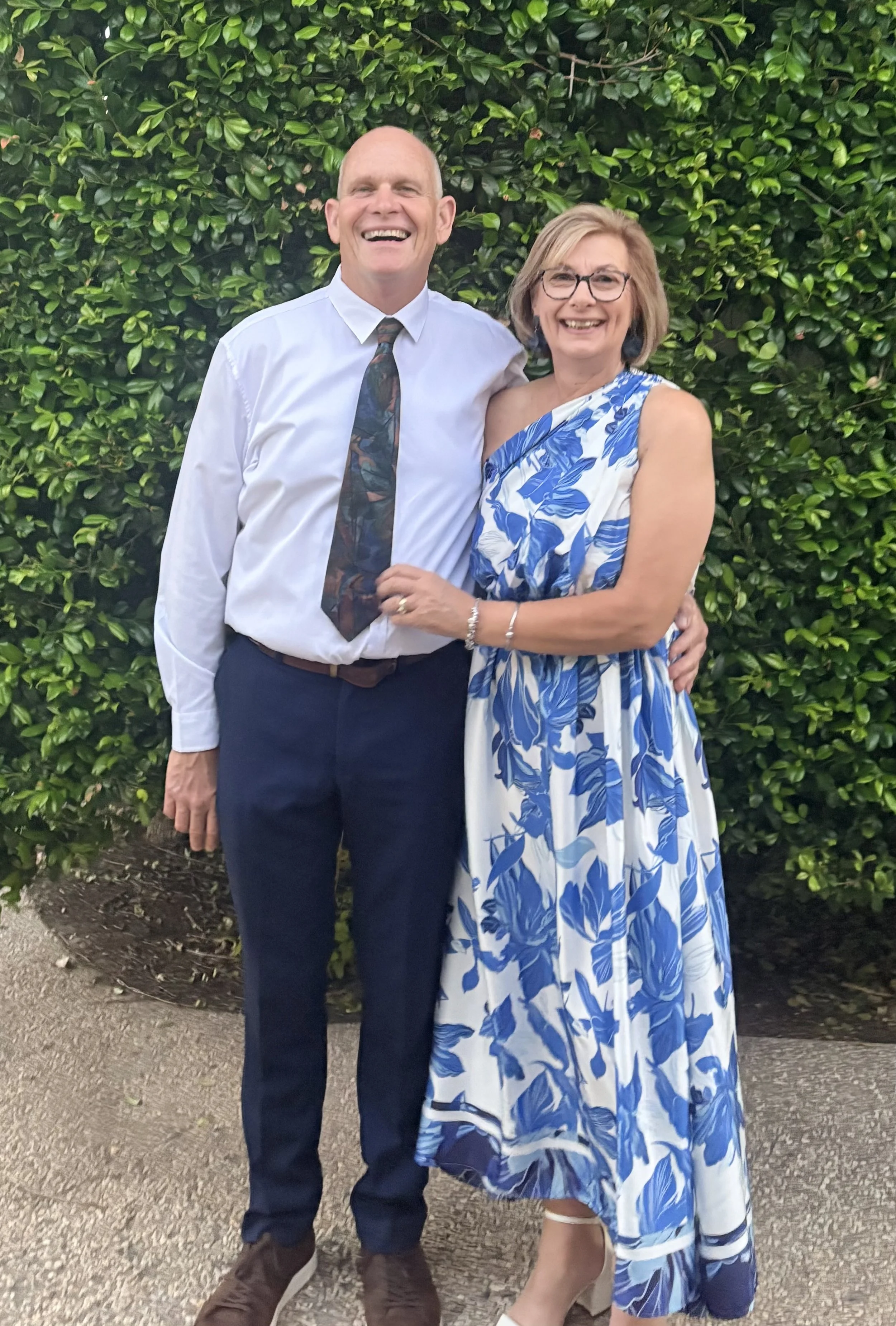 The founders standing close together outdoors in front of green bushes. The man is wearing a white shirt, colorful tie, and dark pants. The woman is wearing glasses and a blue and white floral dress. They appear happy and affectionate.