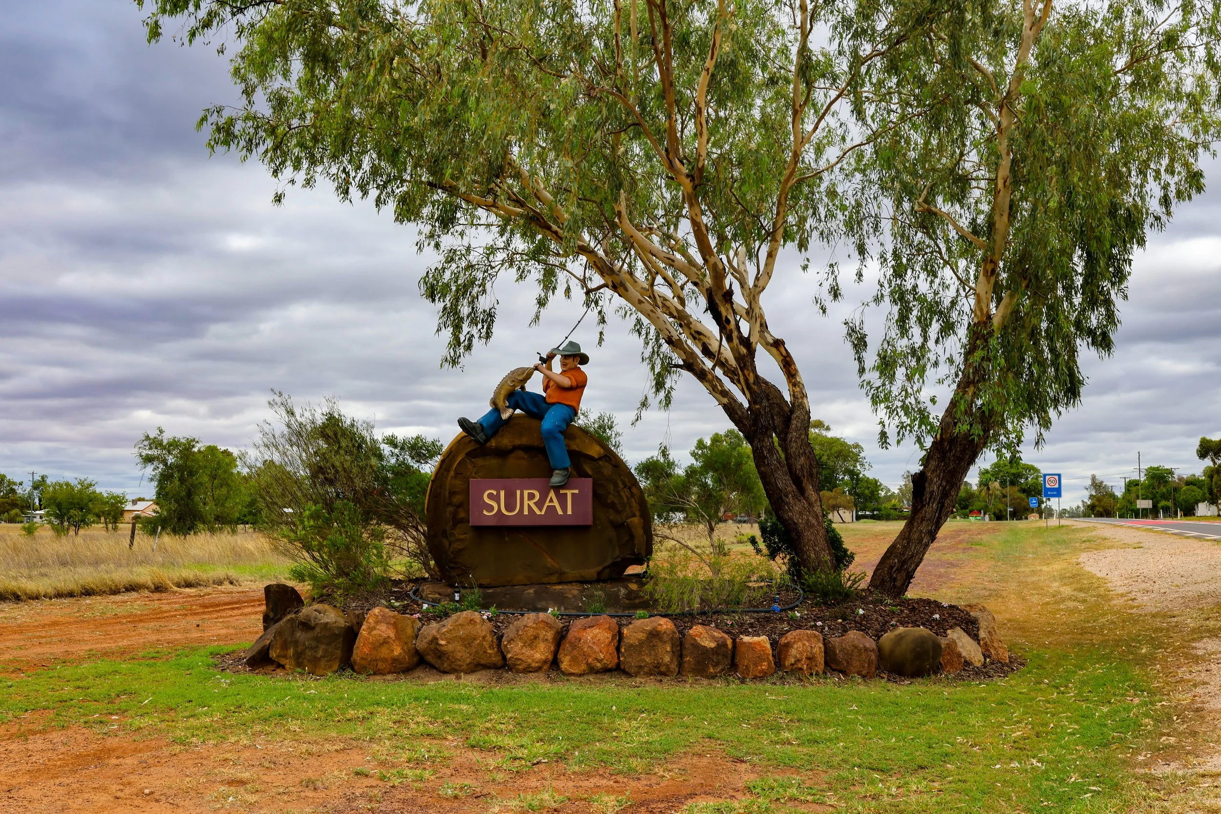 A roadside monument featuring a sculpture of a man in an orange shirt and blue pants sitting on a large wooden log with a sign reading 'SURAT'. The sculpture depicts a man with a hat. The monument is set amidst a landscaped area with rocks and a tree with broad green leaves, under a cloudy sky.