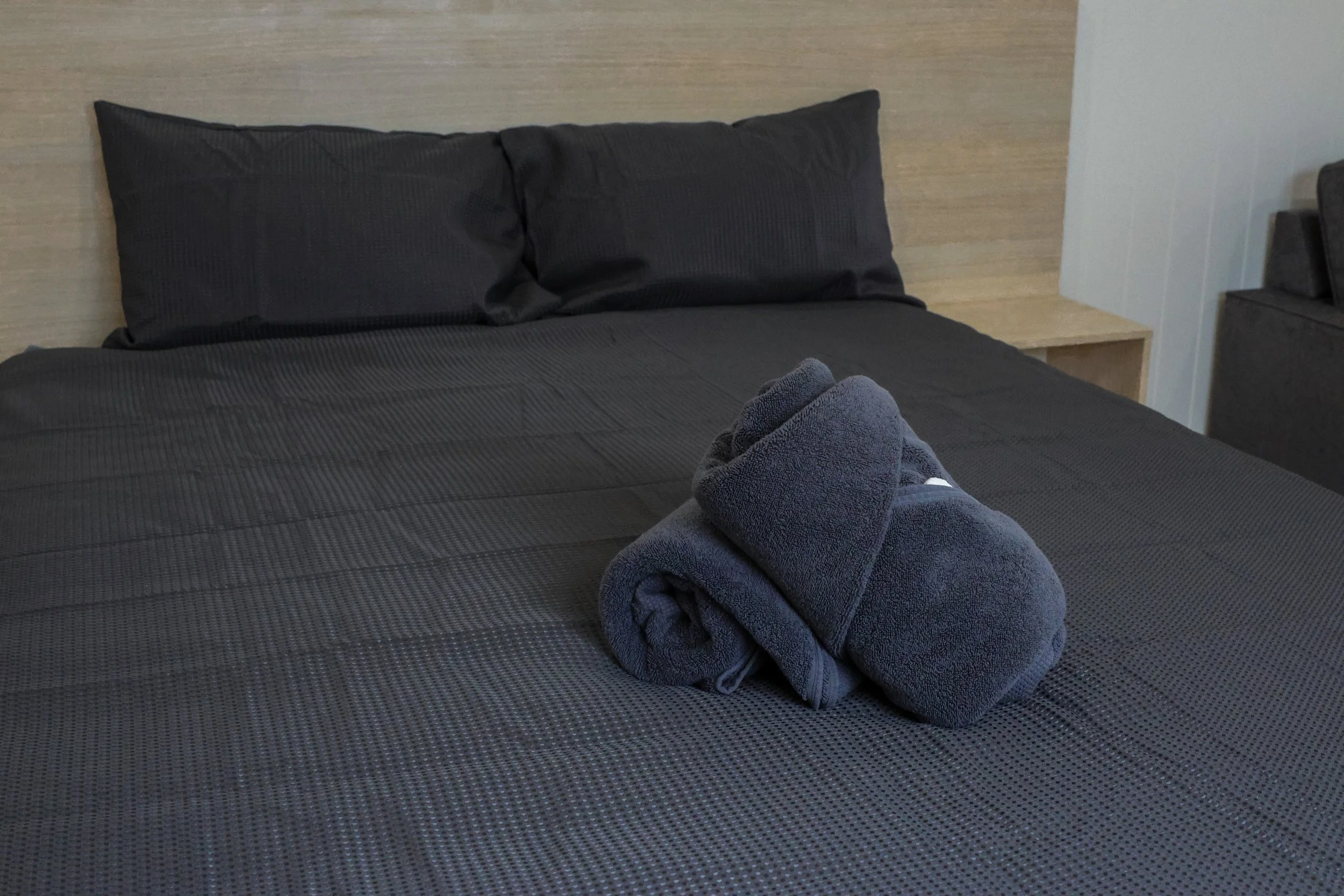 A neatly made bed with black sheets and two black pillows, with a folded dark gray towel on top of the bed.