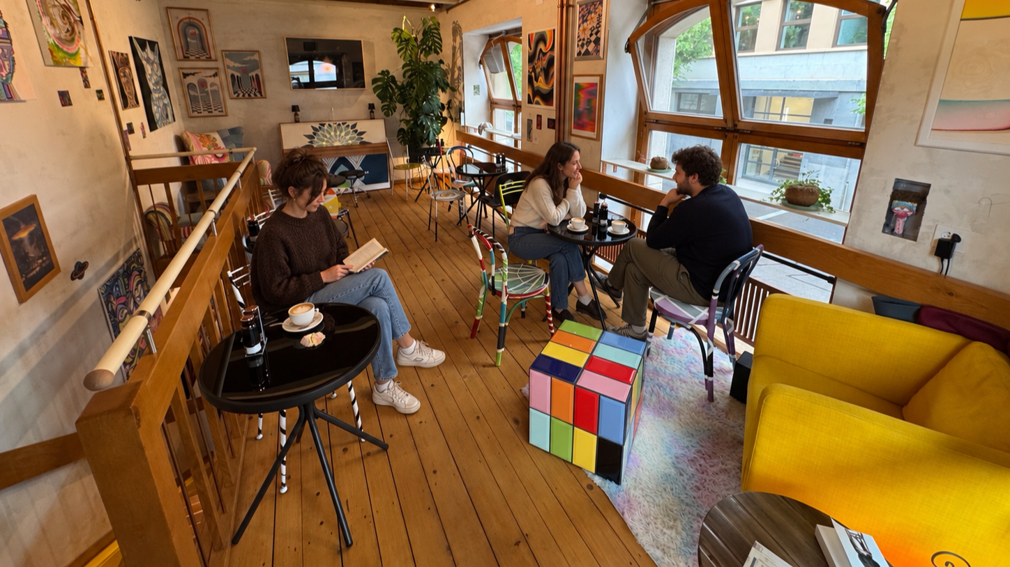 A cozy cafe with three people sitting at separate tables, two women and a man, engaged in conversation and reading. The cafe features wooden floors, colorful artwork on the walls, large windows with a view of neighboring buildings, and vibrant furniture including a yellow sofa and a cube-shaped table with multicolored squares.