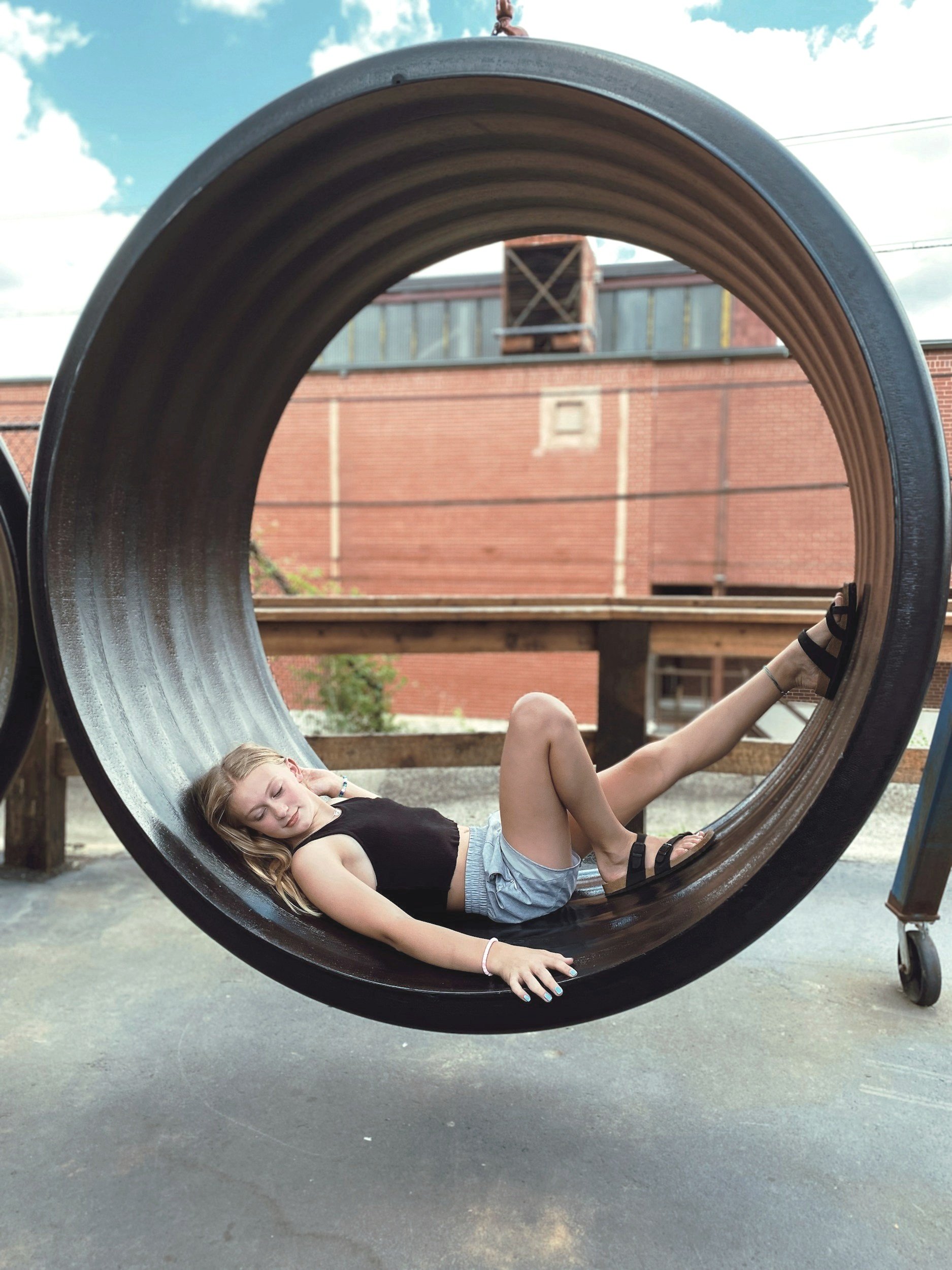 Girl lying inside a large black metal pipe outdoors, with her eyes closed, wearing a black sleeveless top, gray shorts, and sandals.