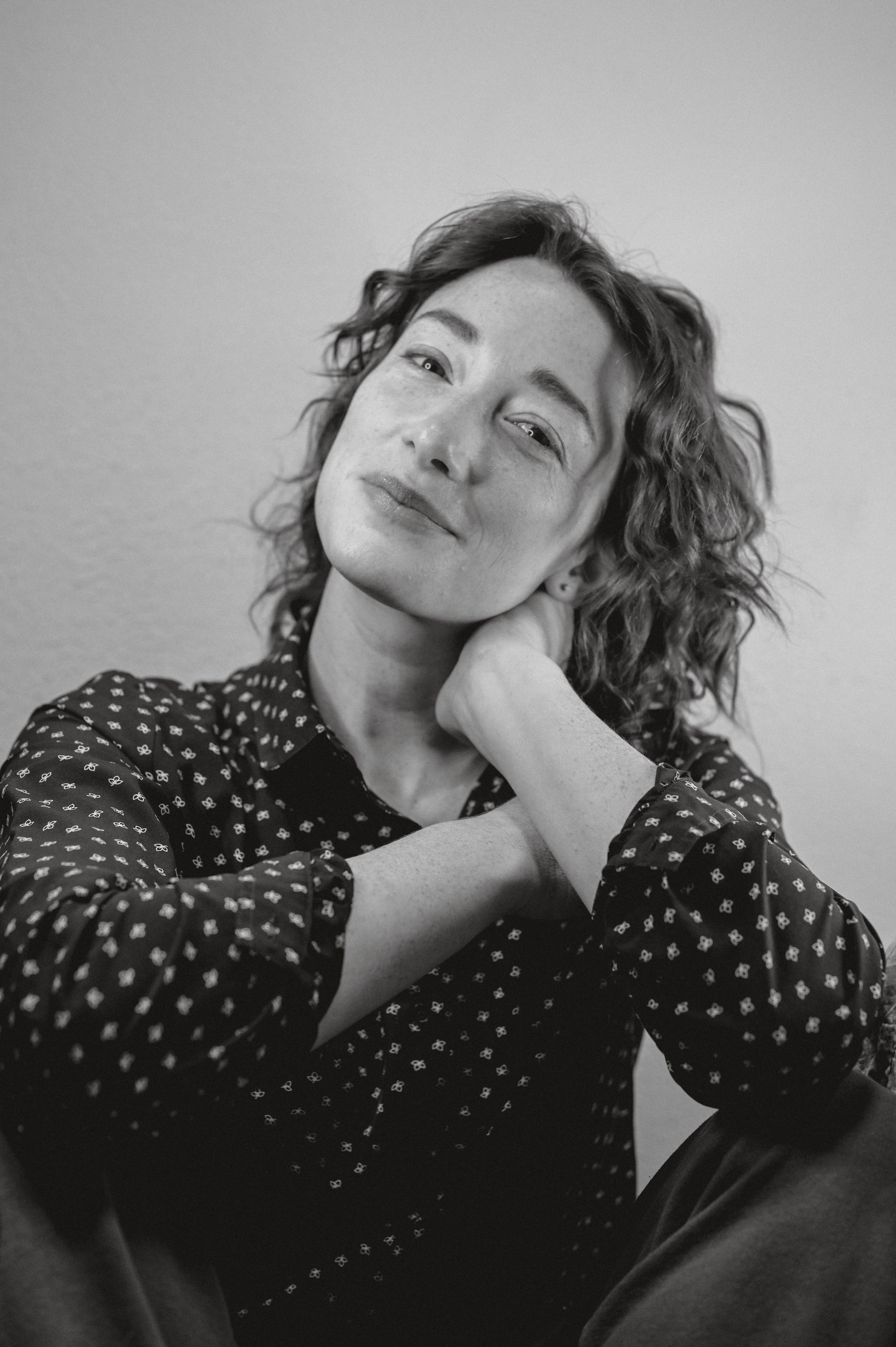Black and white portrait of a woman with curly hair, wearing a patterned blouse, resting her chin on her hand, looking at the camera.