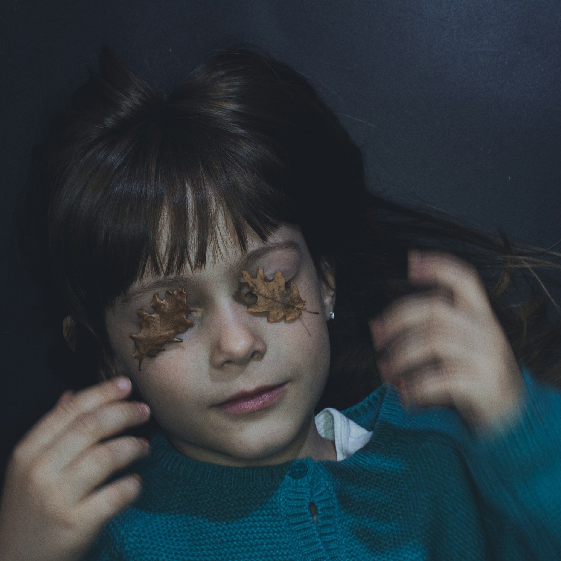 A young girl with long, dark hair lying down with her eyes closed. Two dry autumn leaves cover her eyes, and she is wearing a blue sweater. She appears peaceful and relaxed.