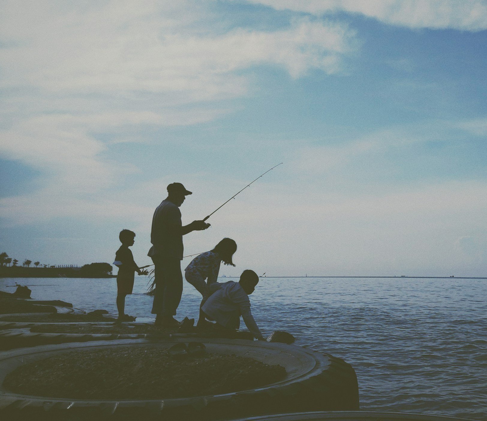 Family fishing together by the water on a partly cloudy day.