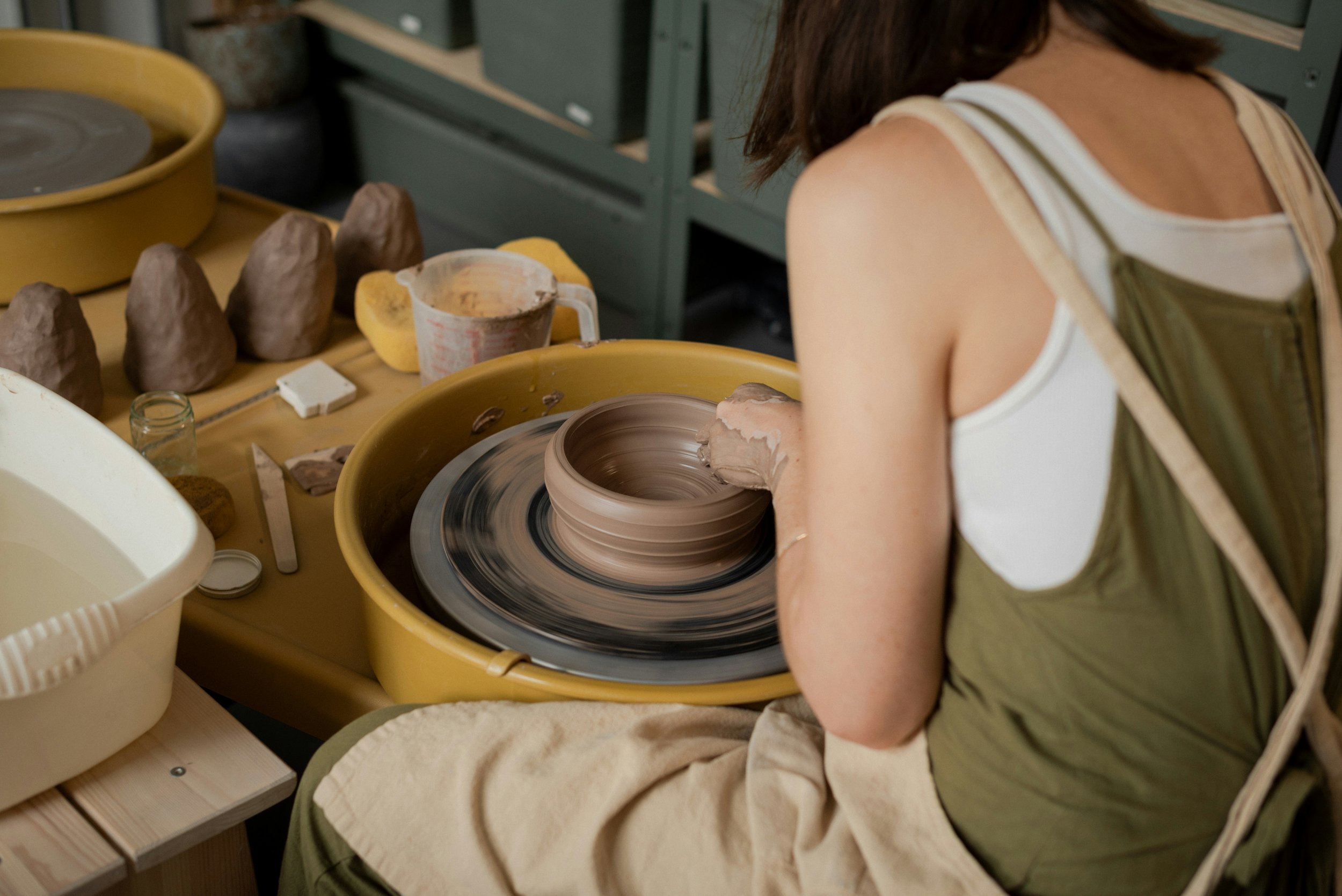 A person shaping clay on a pottery wheel in a studio surrounded by pottery tools and finished clay pieces.