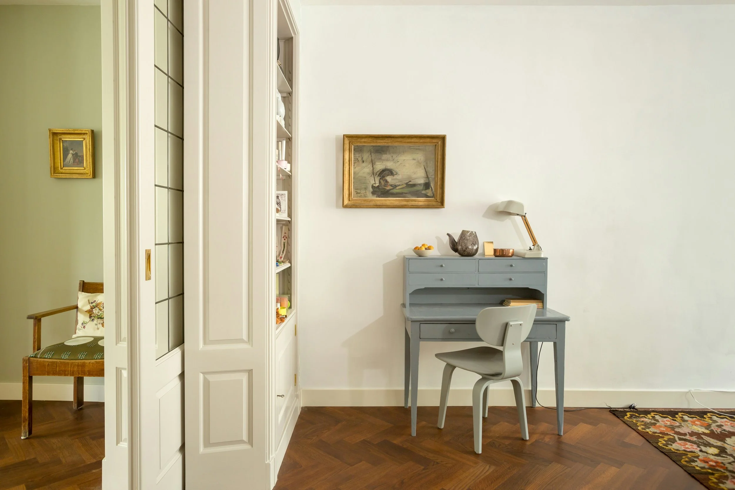 A corner of a room with a light gray writing desk and chair, a white lamp, a framed painting, and some decorative items on the desk. To the left is a white bookshelf, and to the far left is a wooden chair with floral cushion, all on a wooden floor.