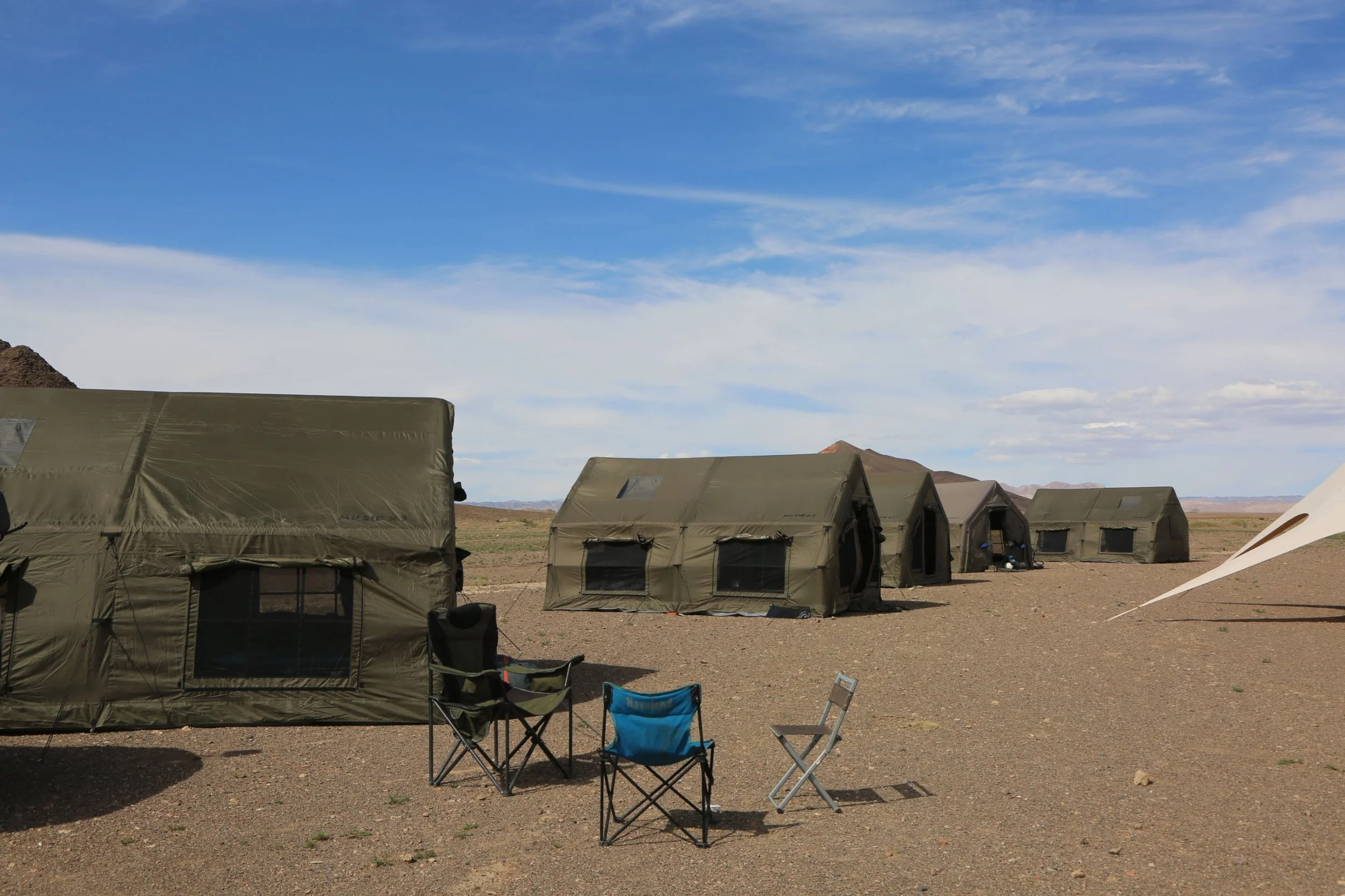 Military tents set up in a barren desert landscape under a partly cloudy blue sky, with mountain ranges in the background and camping chairs in the foreground.