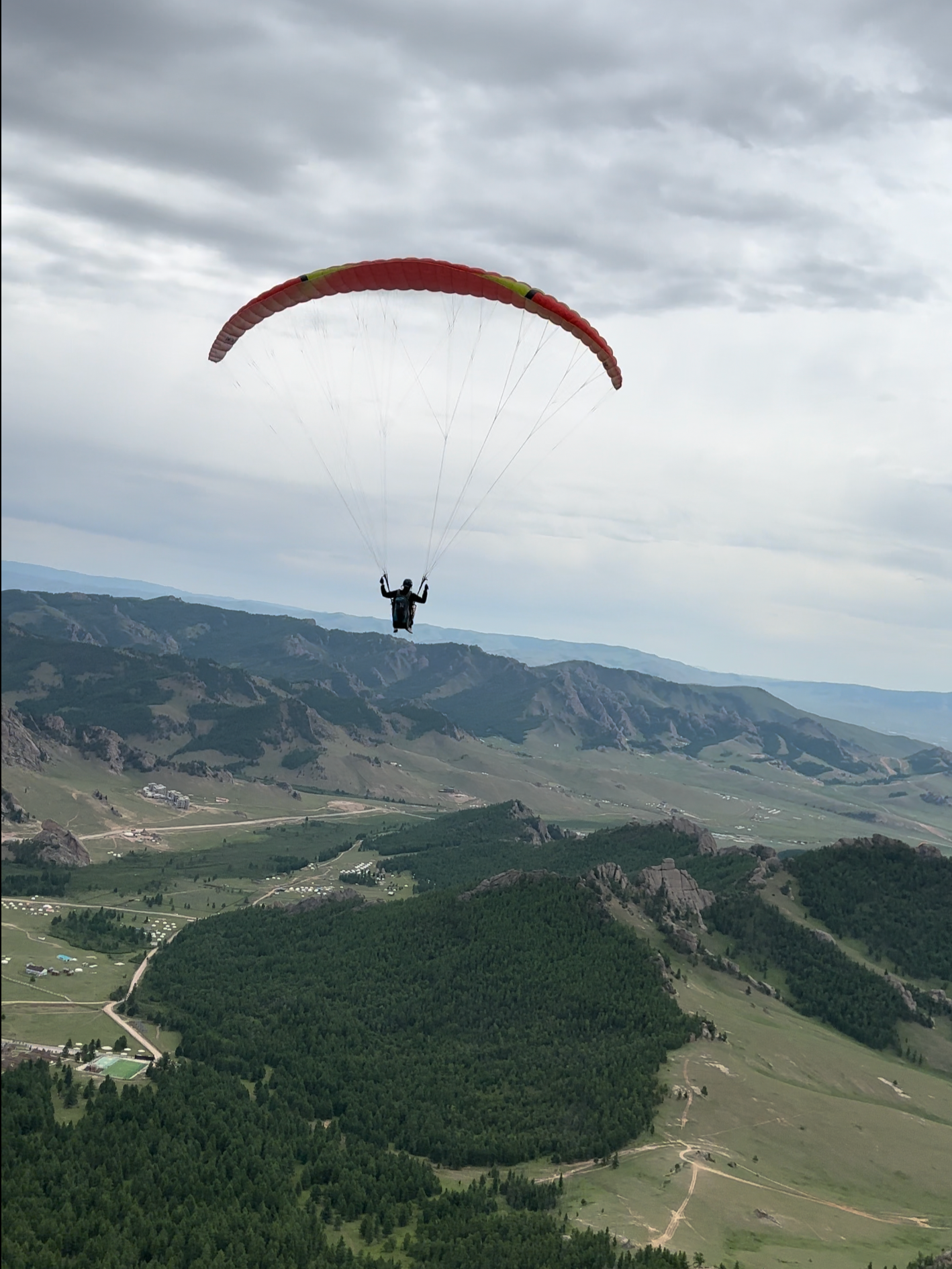 Paraglider flying over a lush green valley with mountains in the background on a cloudy day.