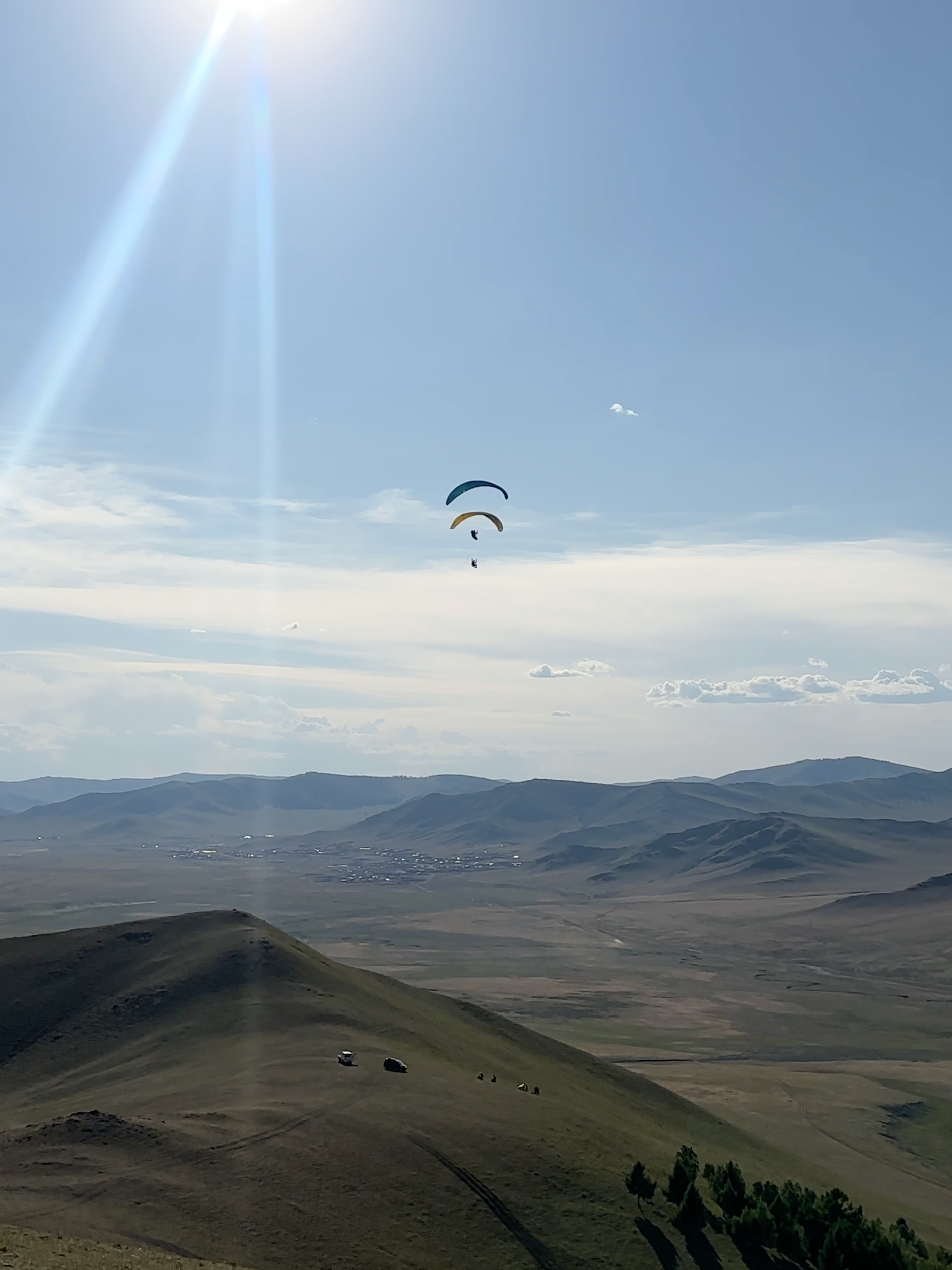 Two paragliders soaring above a landscape of rolling hills and mountains on a clear, sunny day with a bright sun and blue sky.