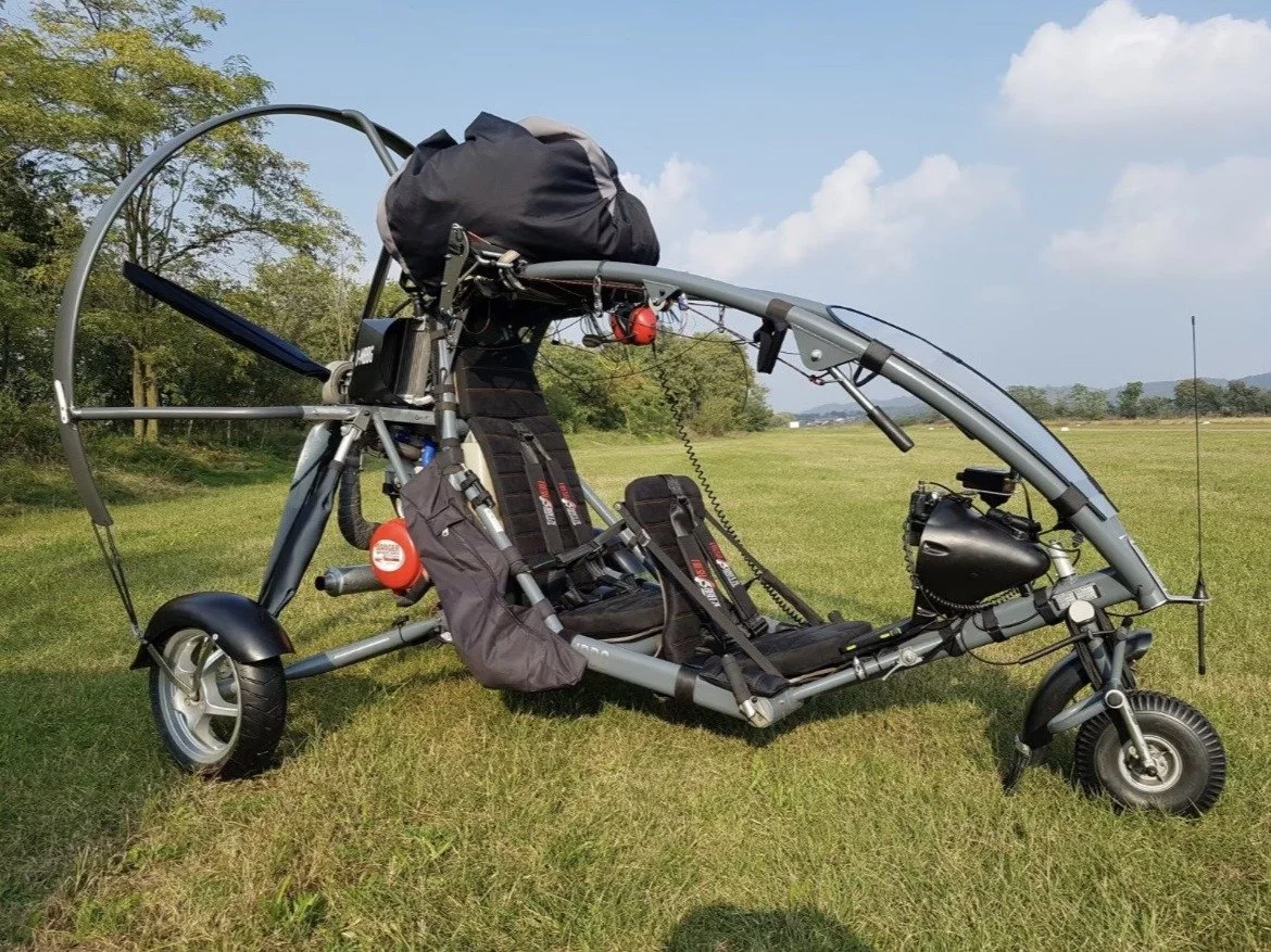 Two-seat tandem paramotor trike with its wing packed on top, parked on a grass airfield