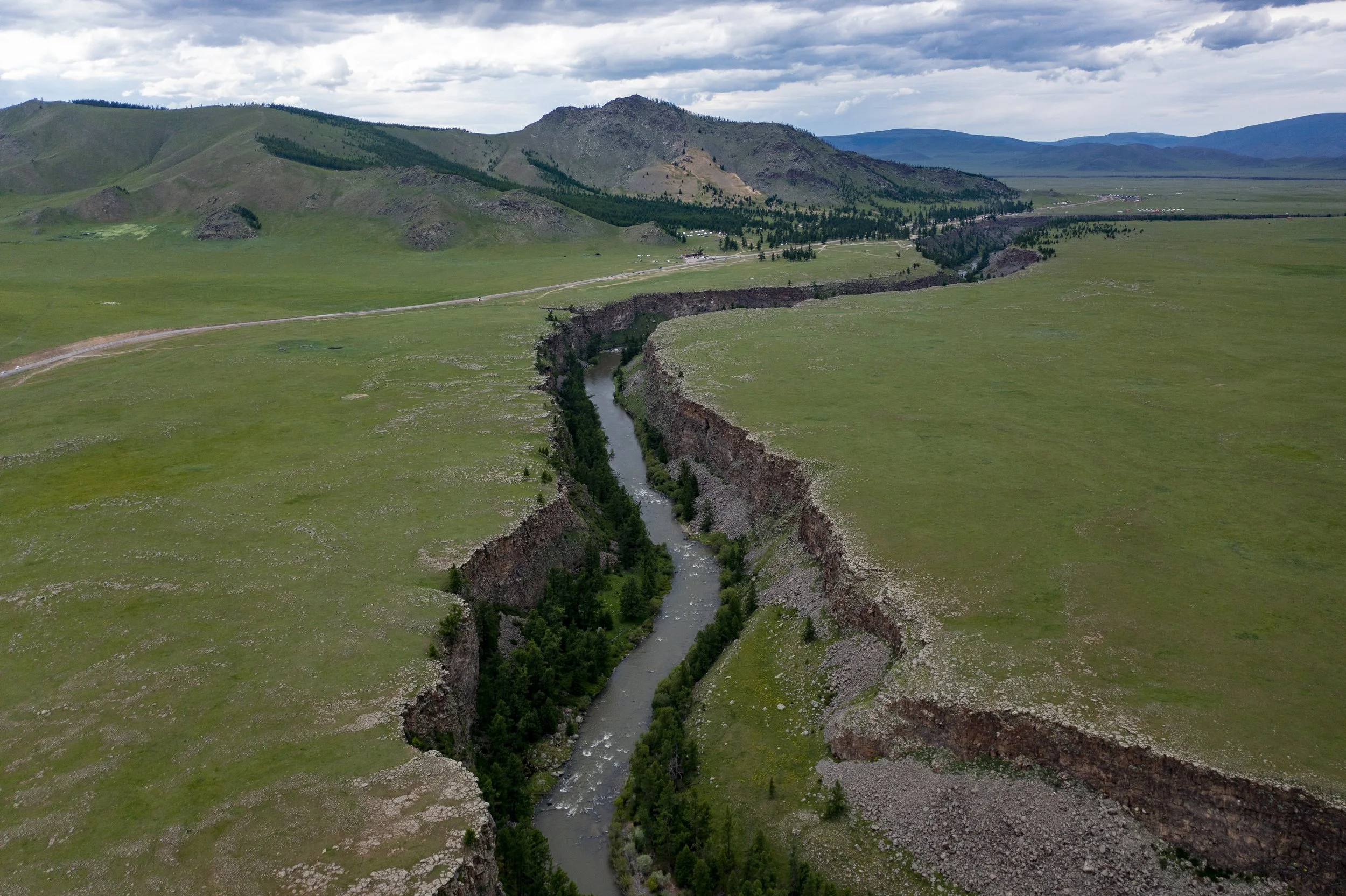 Aerial view of a deep river canyon with green vegetation along the riverbanks, surrounded by rolling green hills and distant mountains under a partly cloudy sky.