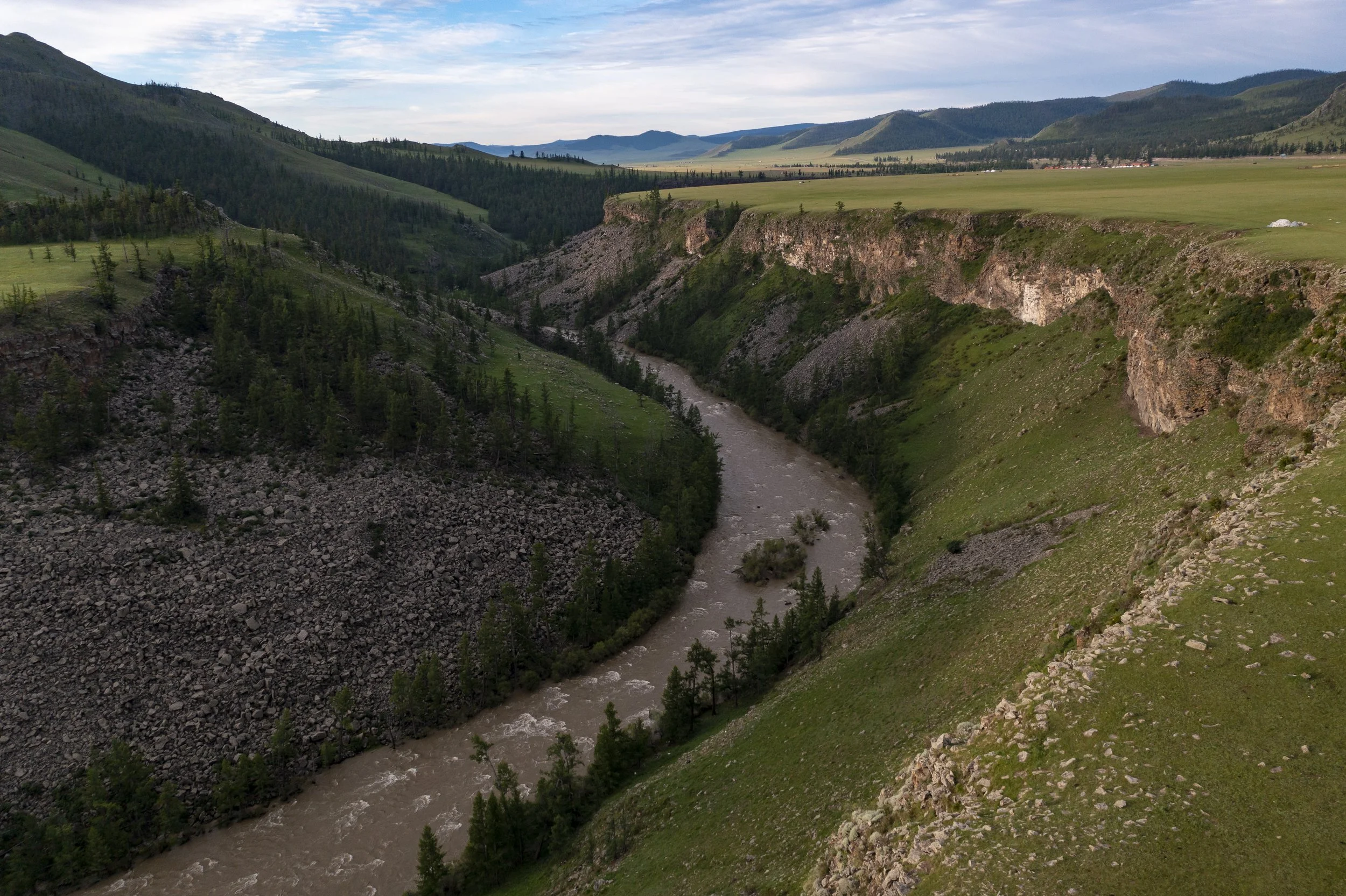 A river flowing through a green canyon with steep, rocky cliffs, surrounded by hills and mountains under a partly cloudy sky.