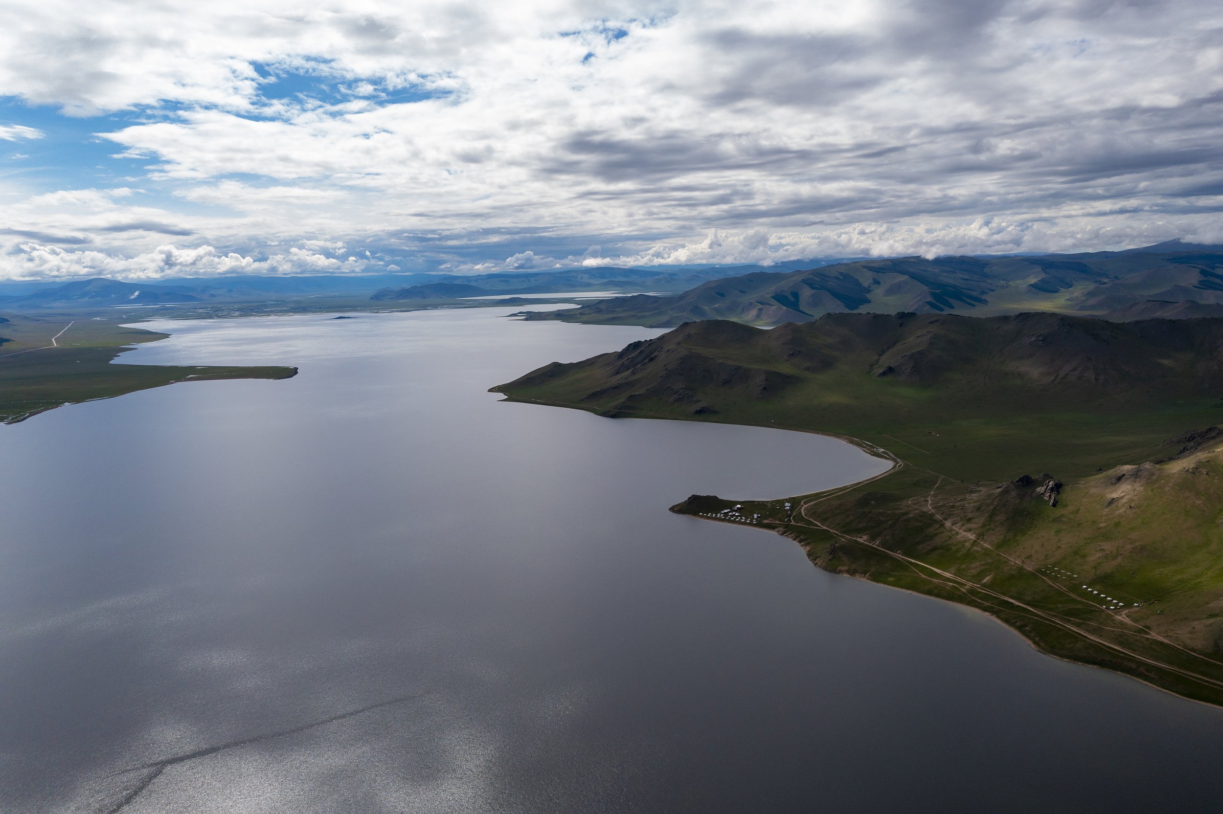 Aerial landscape of a large lake surrounded by green hills and mountains under a partly cloudy sky.