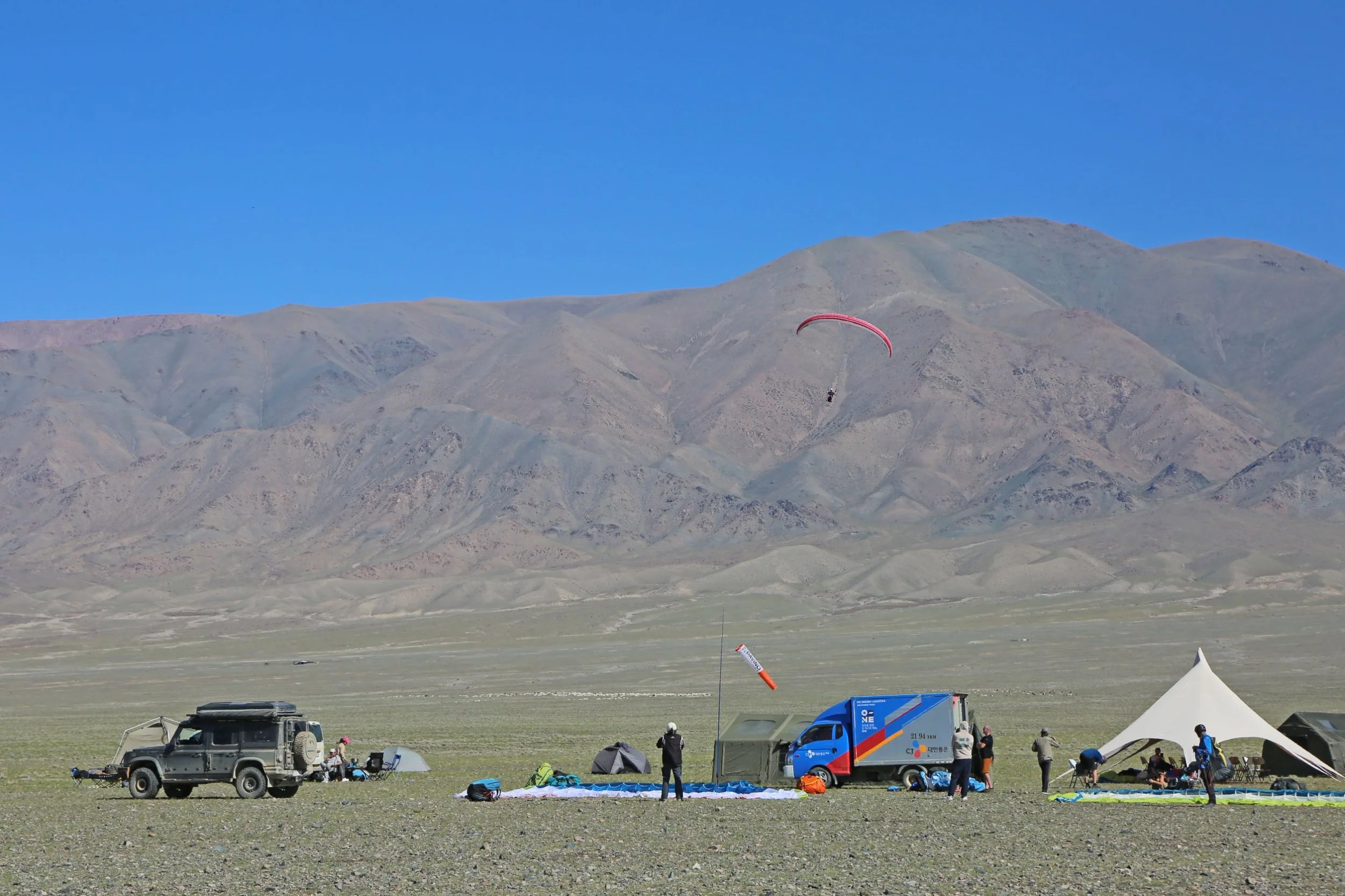 People preparing for a paragliding flight on a grassy flat area with tents, equipment, and vehicles, set against a backdrop of mountains and a clear blue sky.