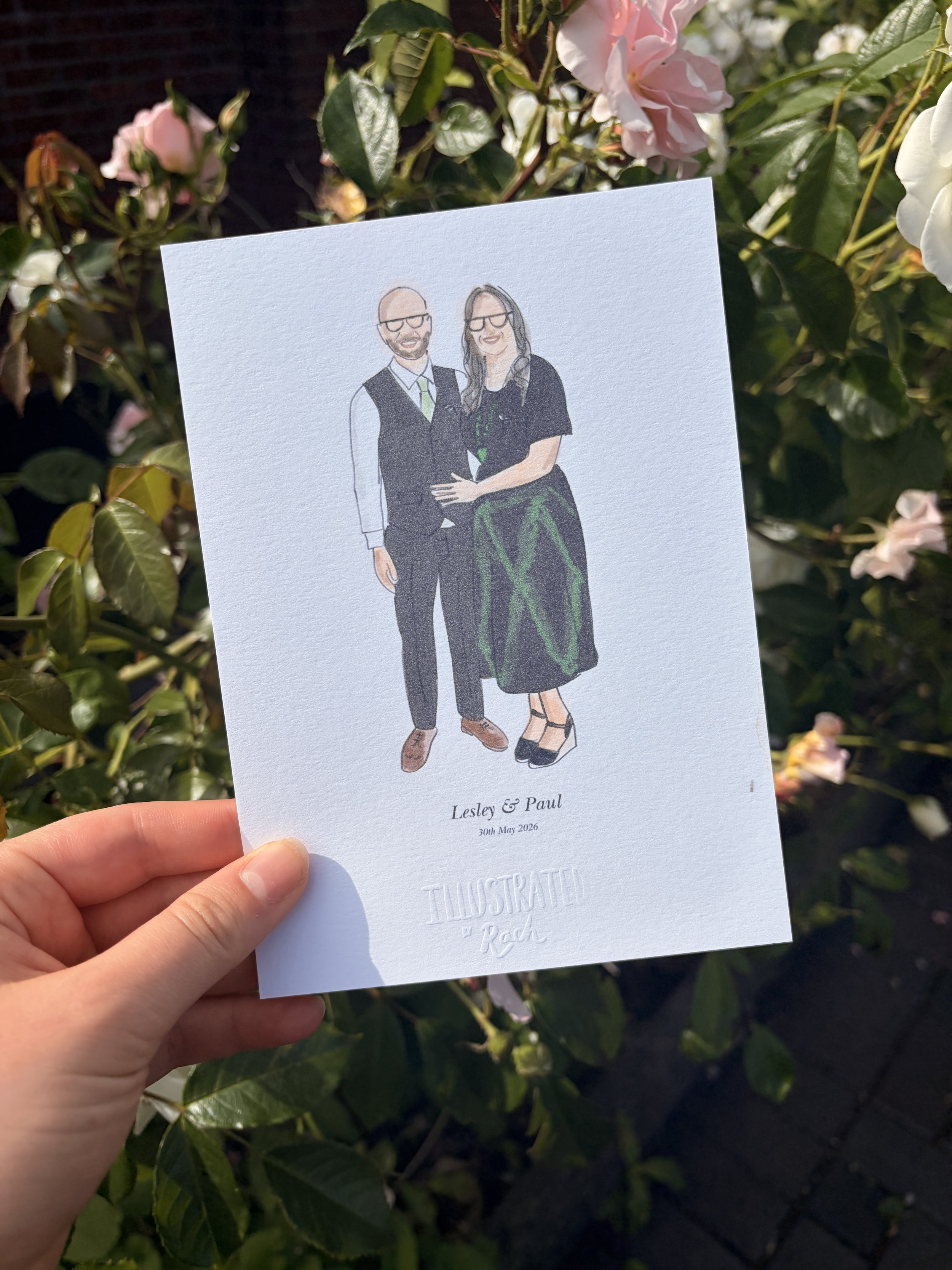 A hand holding a wedding invitation card with an illustration of a couple in front of greenery and flowers, under sunlight, with the names Lesley and Paul and the date 30th May 2026.