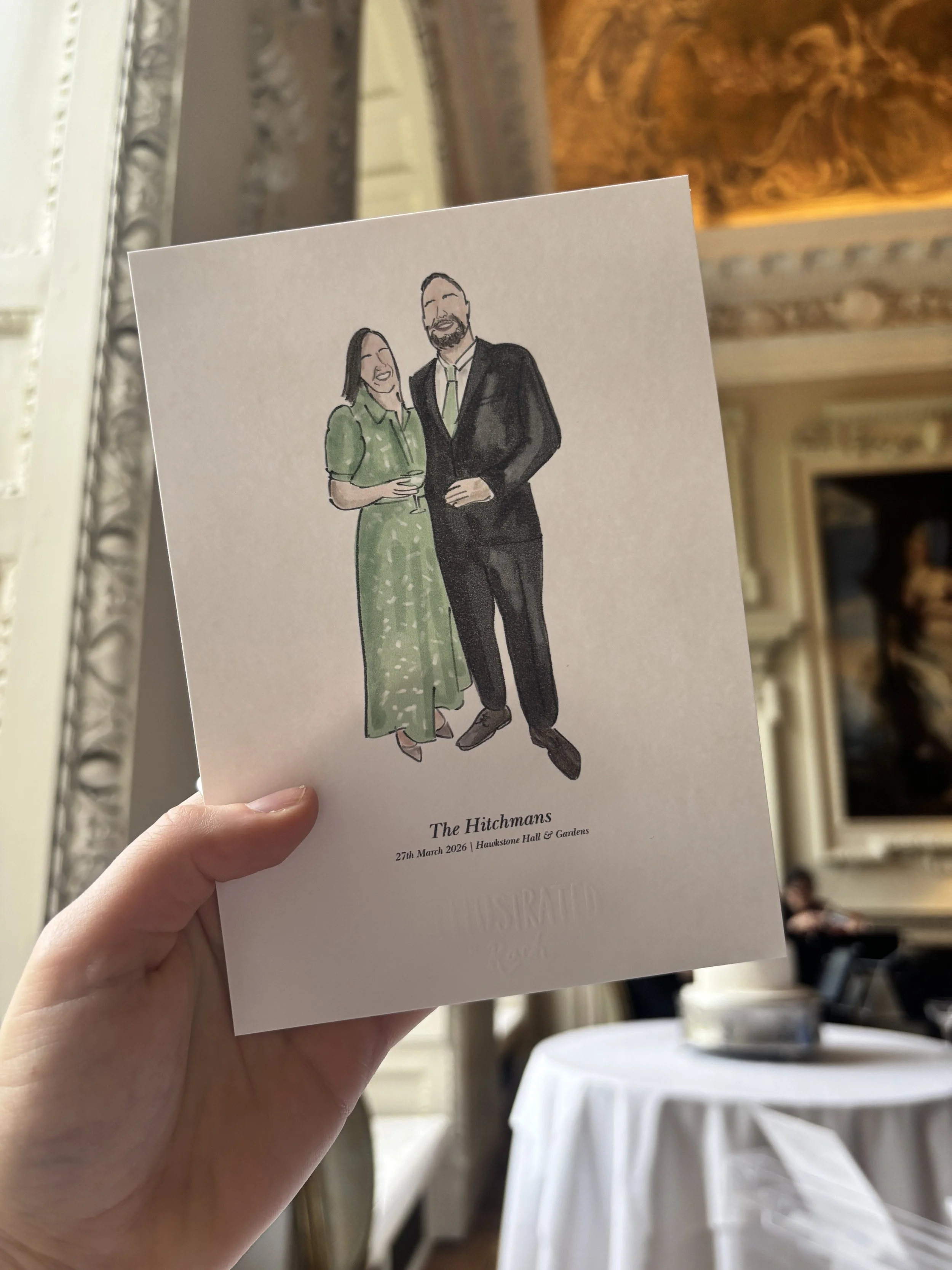 A hand holding a printed illustration of a man and woman dressed in formal attire, standing close and smiling. The text below the illustration reads: "The Hitchmans, 27th March 2026, Hawkstone Hall & Gardens." In the background, there is a decorative