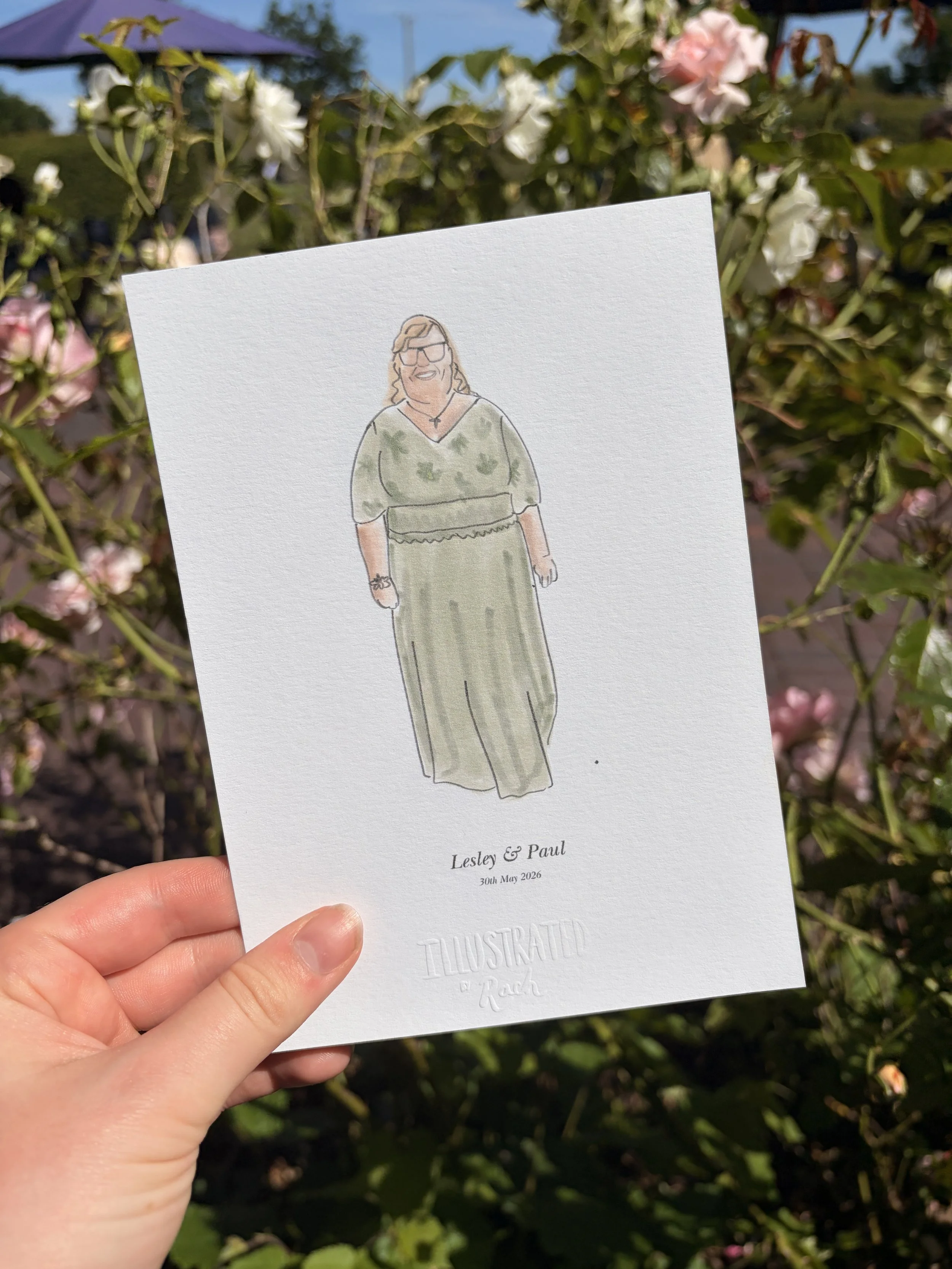 Hand holding a wedding invitation with an illustration of a woman in a green dress and sunglasses, set outdoors amidst blooming roses.