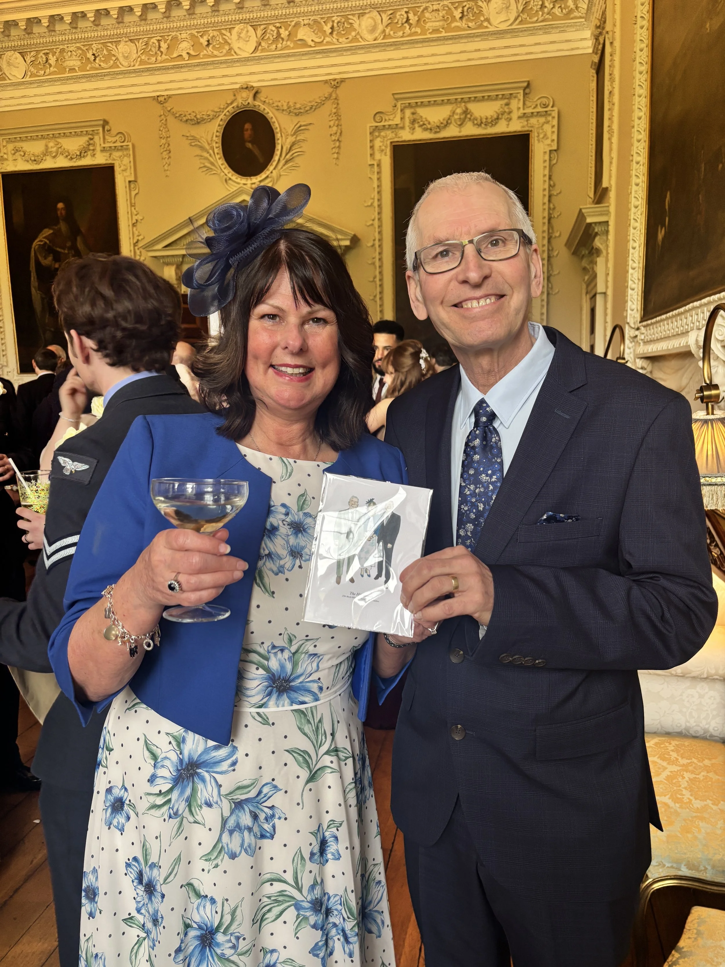 A man and woman smiling at a formal event in an ornate room with gold accents and paintings. The woman is holding a drink and a paper, and the man is holding a piece of paper.