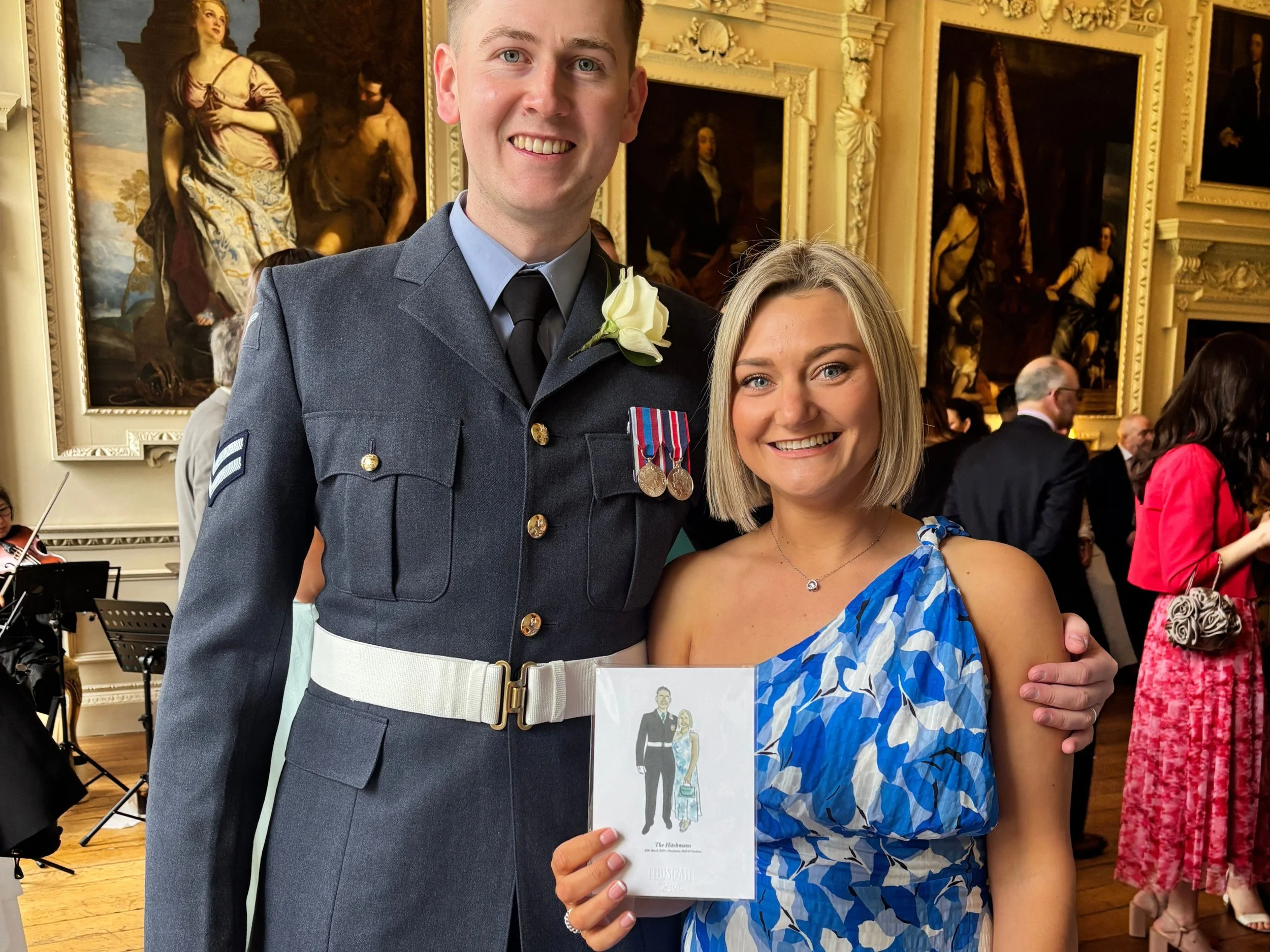 A smiling man in a military uniform with medals standing next to a woman in a blue patterned dress at an art gallery event. The woman is holding a program or booklet.