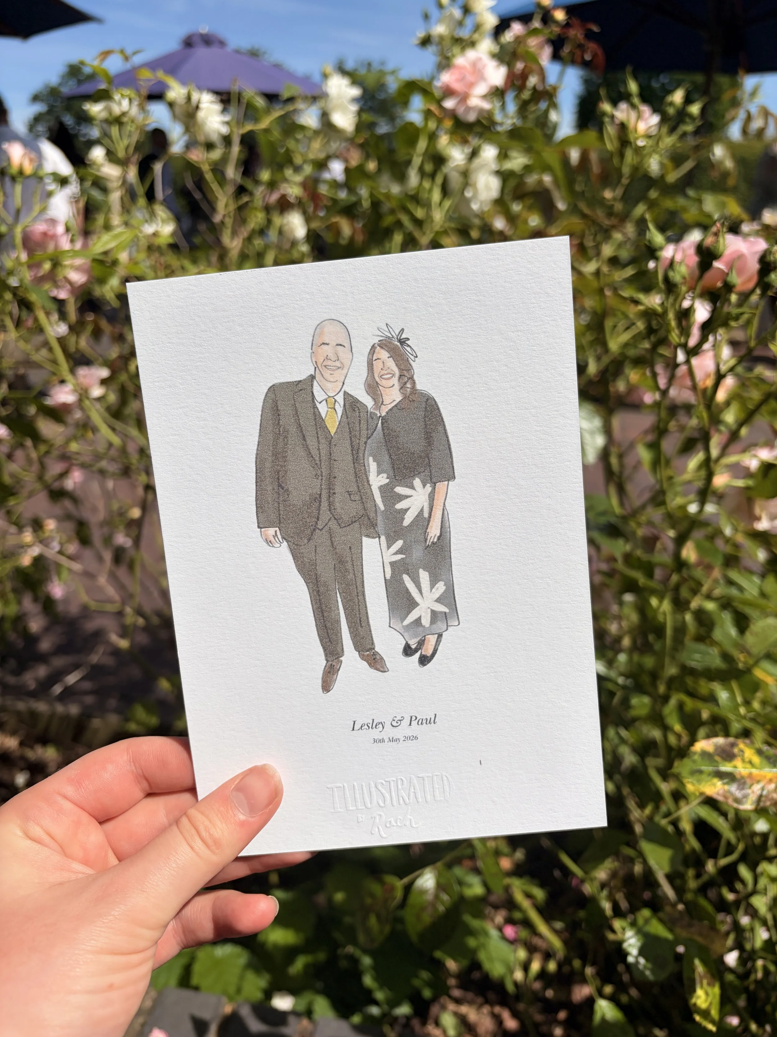 A hand holding a wedding invitation card with illustrated couple, Lesley and Paul, on a garden background with pink and white flowers.