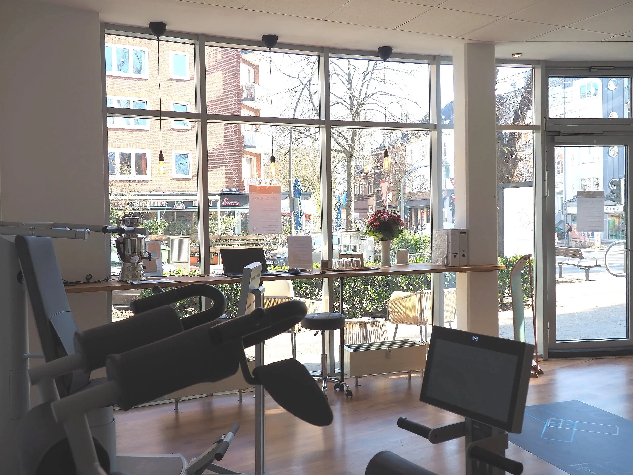 Innenraum eines Fitnessstudios mit Trainingsgeräten, Blick nach draußen durch große Fenster auf Straßen und Gebäude, Tageslicht