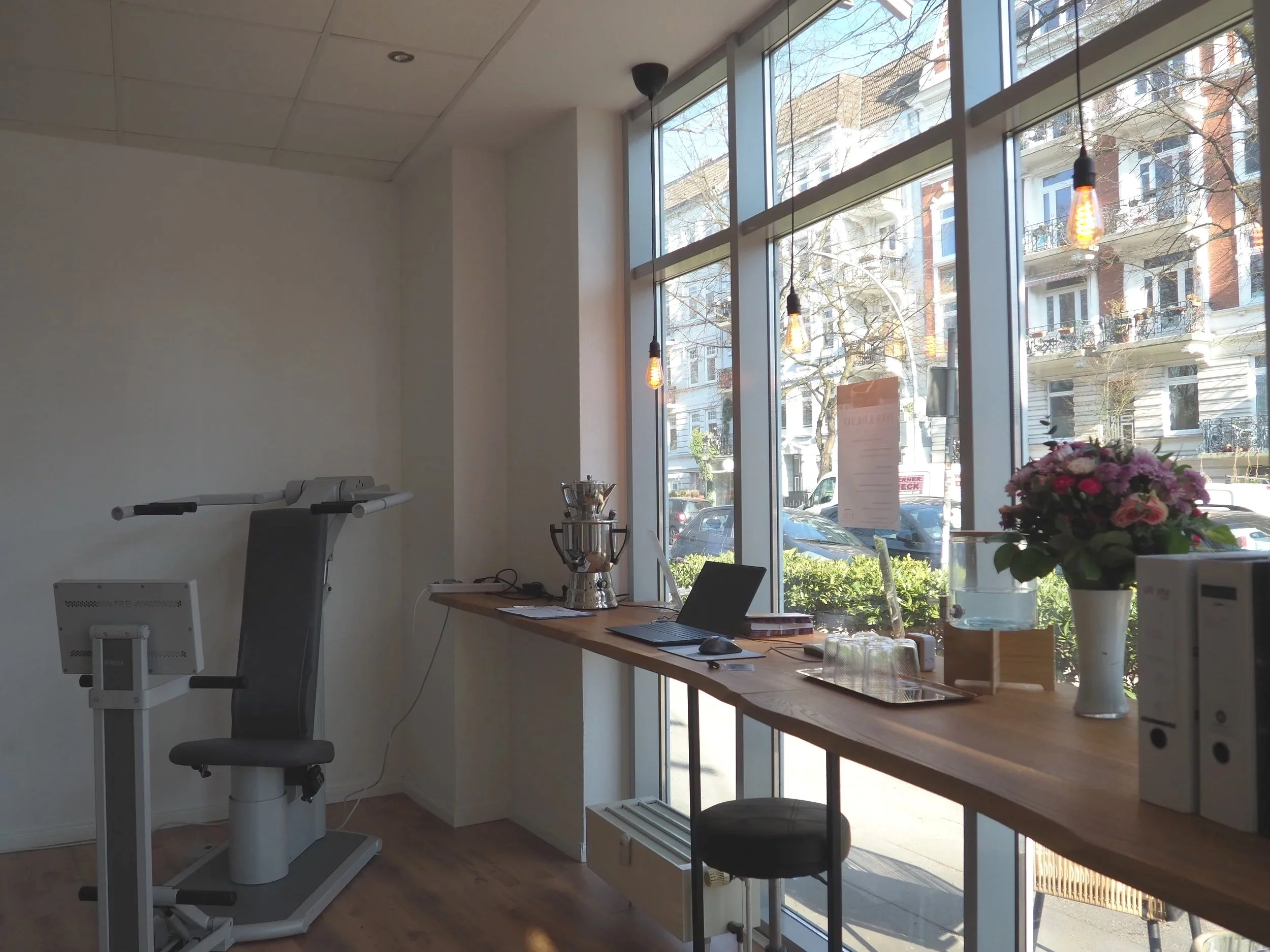 Inneres eines gemütlichen Cafés mit großen Fenster, einem Laptop, einem Blumenstrauß, und einigen Trophäen auf einem Holzregal, abends mit warmem Licht.