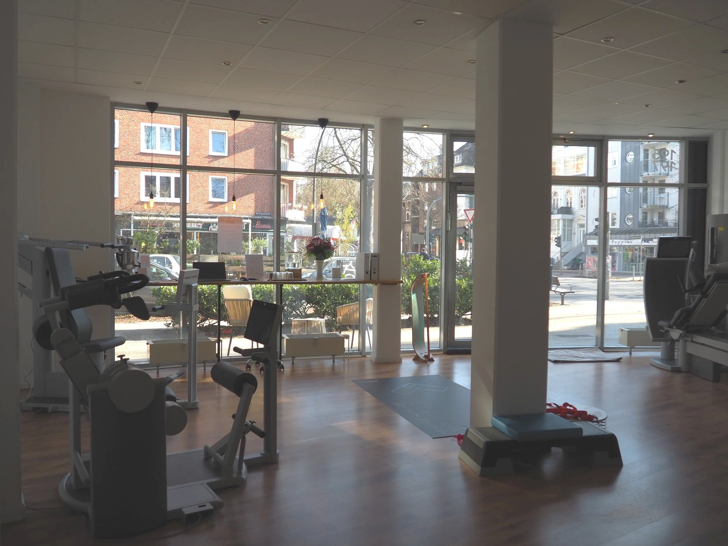 Innenansicht eines Fitnessstudios mit Fitnessgeräten, einem Stehständer, einer Yogamatte, einem Skateboard, und großen Fenstern, die einen Blick auf die Straße und schöne Gebäudefassaden bieten.