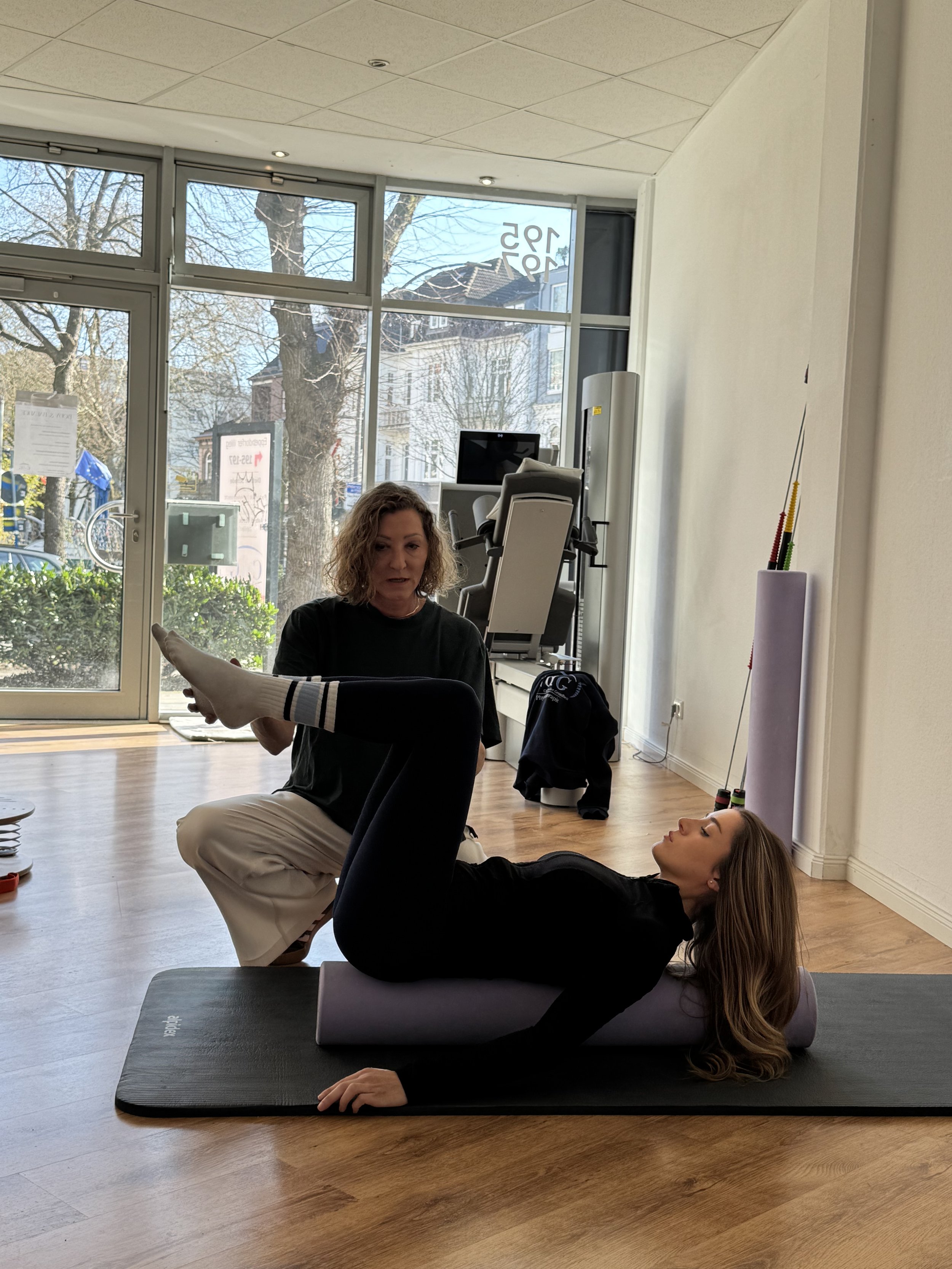 Frau liegt auf Yogamatte, g gelöscht, während eine Trainerin sie im Fitnessstudio anleitet.