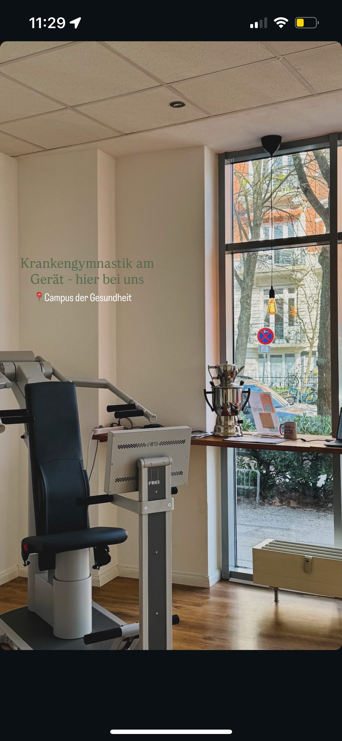 Ein Raum mit medizinischem Gerät, einer schwarzen Sportlehne, einem Schreibtisch mit einer Kaffeemaschine, einem Computer und einem Fenster mit Blick auf Bäume und ein Gebäude.